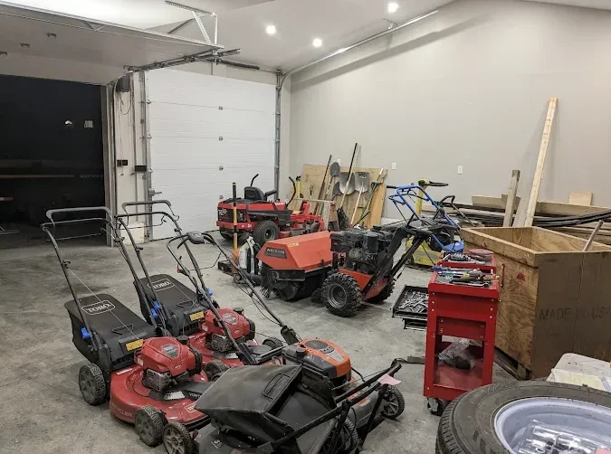 Garage with various lawn equipment, tools, and wooden boards stored inside, including push mowers, a tiller, and a red tool cabinet.