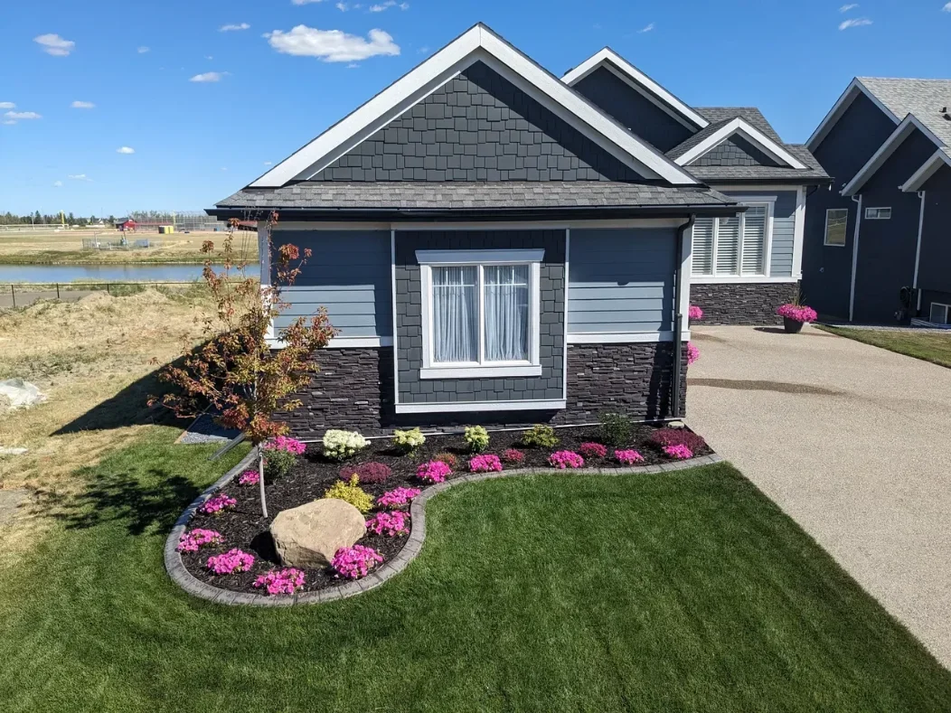 Modern house with landscaped front yard featuring green grass, stone-bordered flower beds, and small shrubs. Clear blue sky in the background.