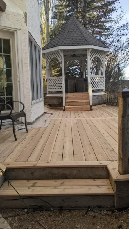 A newly built wooden deck with steps leading up to a white gazebo with a shingled roof, located next to a white house with a window and a black outdoor chair.