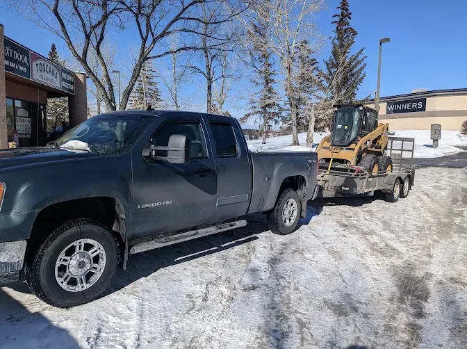 A black pickup truck hauling a trailer with a small backhoe loader on it, parked on a snowy lot outside a shopping center.