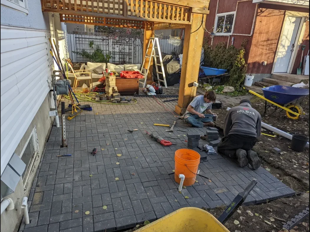 Two workers installing pavers on a backyard patio under a wooden pergola. One worker is kneeling while others sit nearby, surrounded by tools, bricks, and construction materials. A wheelbarrow, ladder, and various outdoor furniture are also visible.