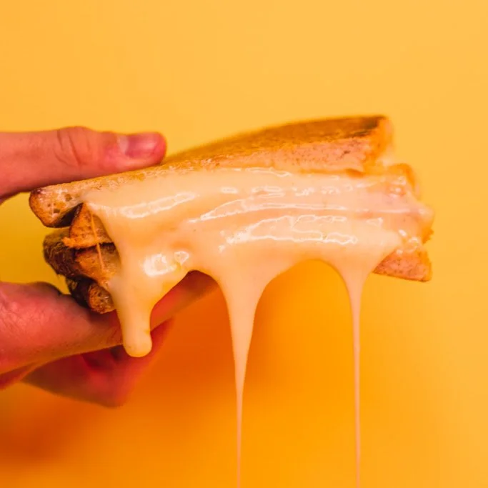 A hand holding a grilled cheese sandwich with melted cheese dripping down the sides against a yellow background.