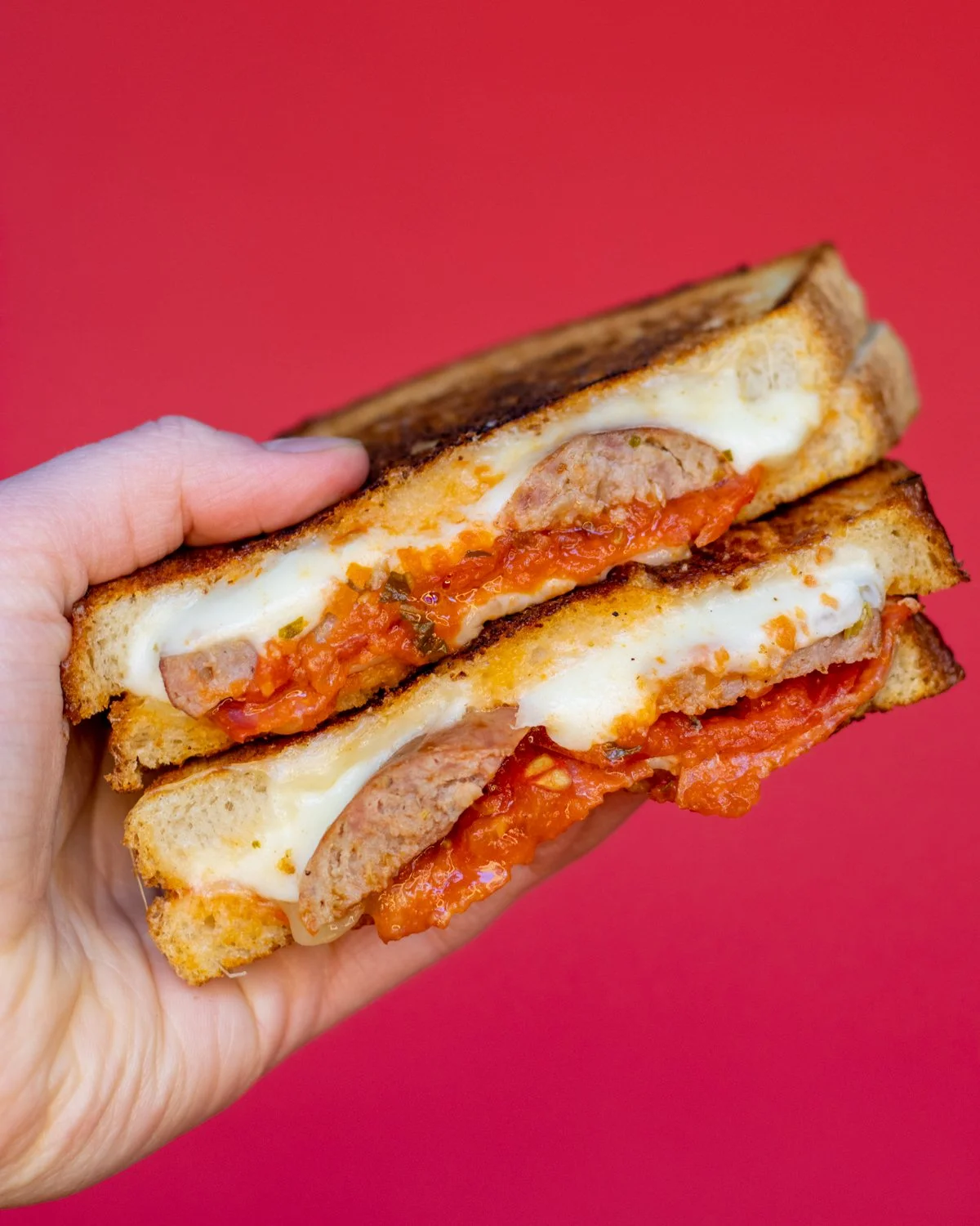 Hand holding a grilled sandwich with sausage, melted cheese, tomato sauce, and bread against a red background.