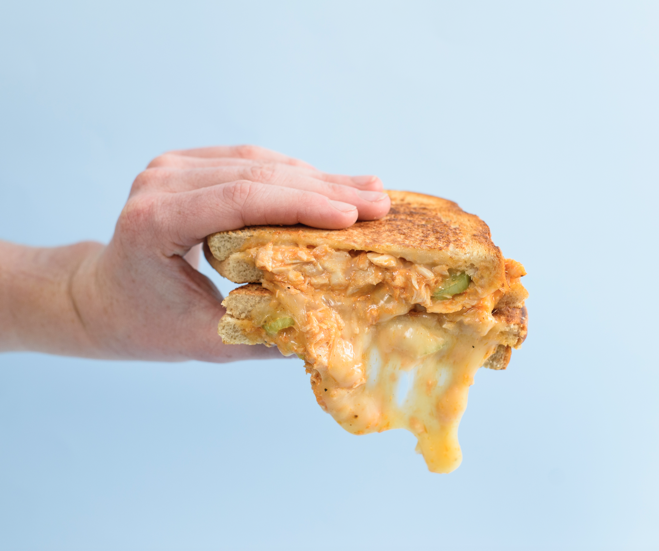 A hand holding a half-eaten grilled cheese sandwich with melted cheese and some vegetables against a light blue background.