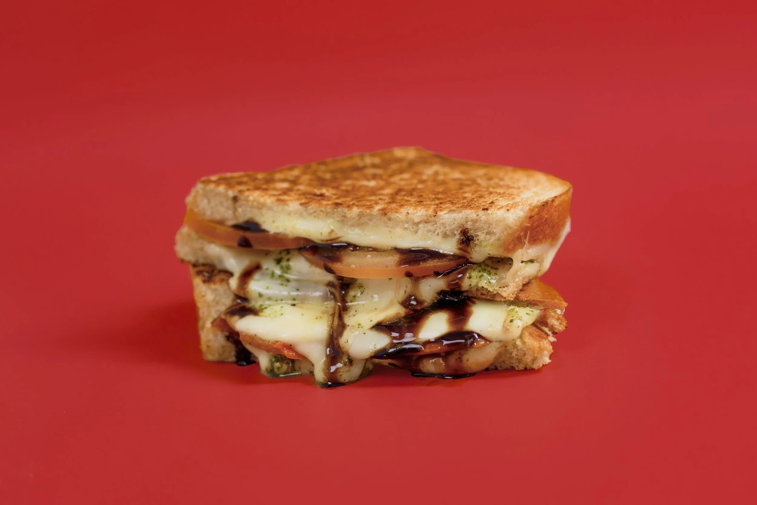 Close-up of a grilled cheese sandwich with melted cheese, tomato slices, and balsamic glaze on red background.