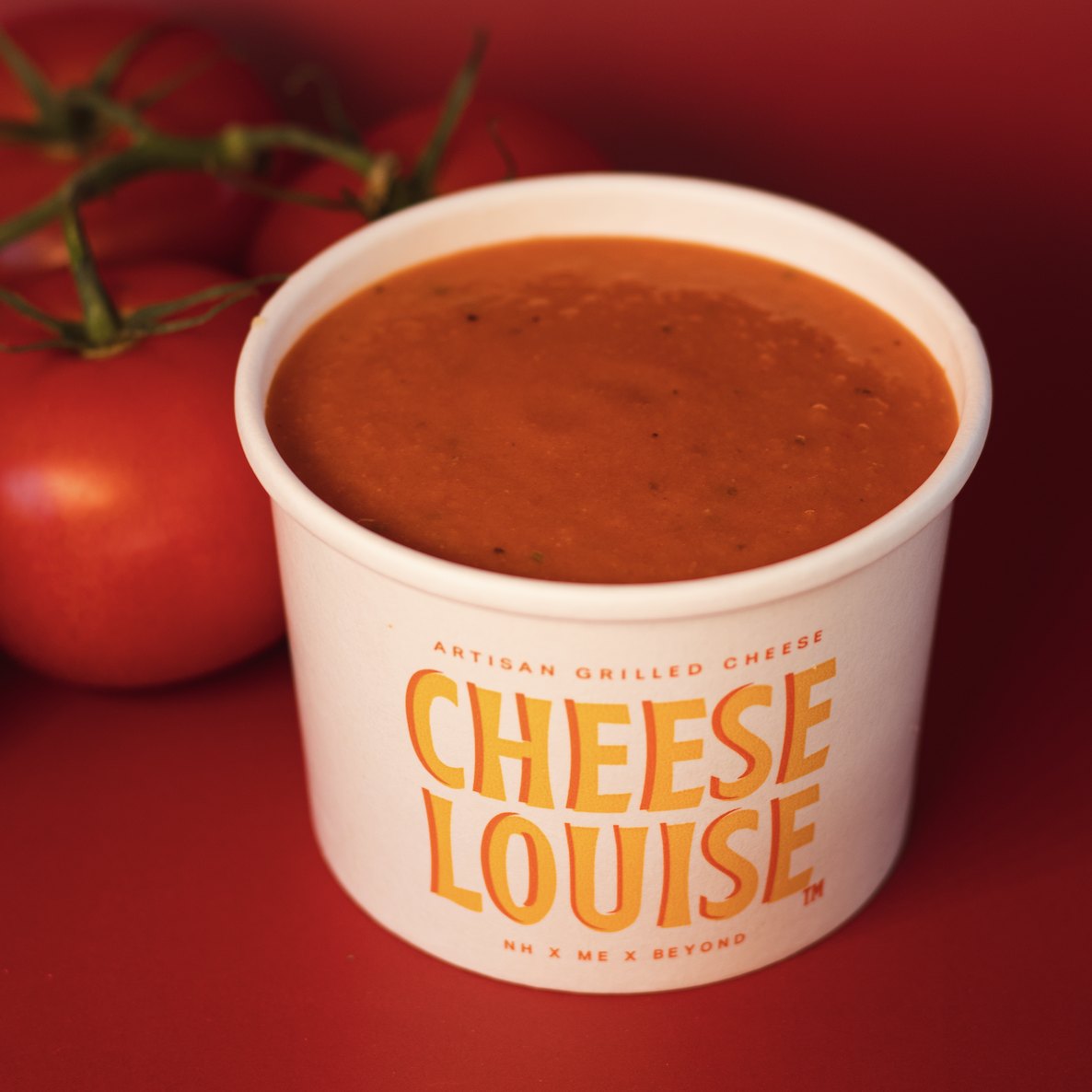 A cup of tomato soup with a label reading 'Cheese Louise' and some fresh tomatoes in the background.