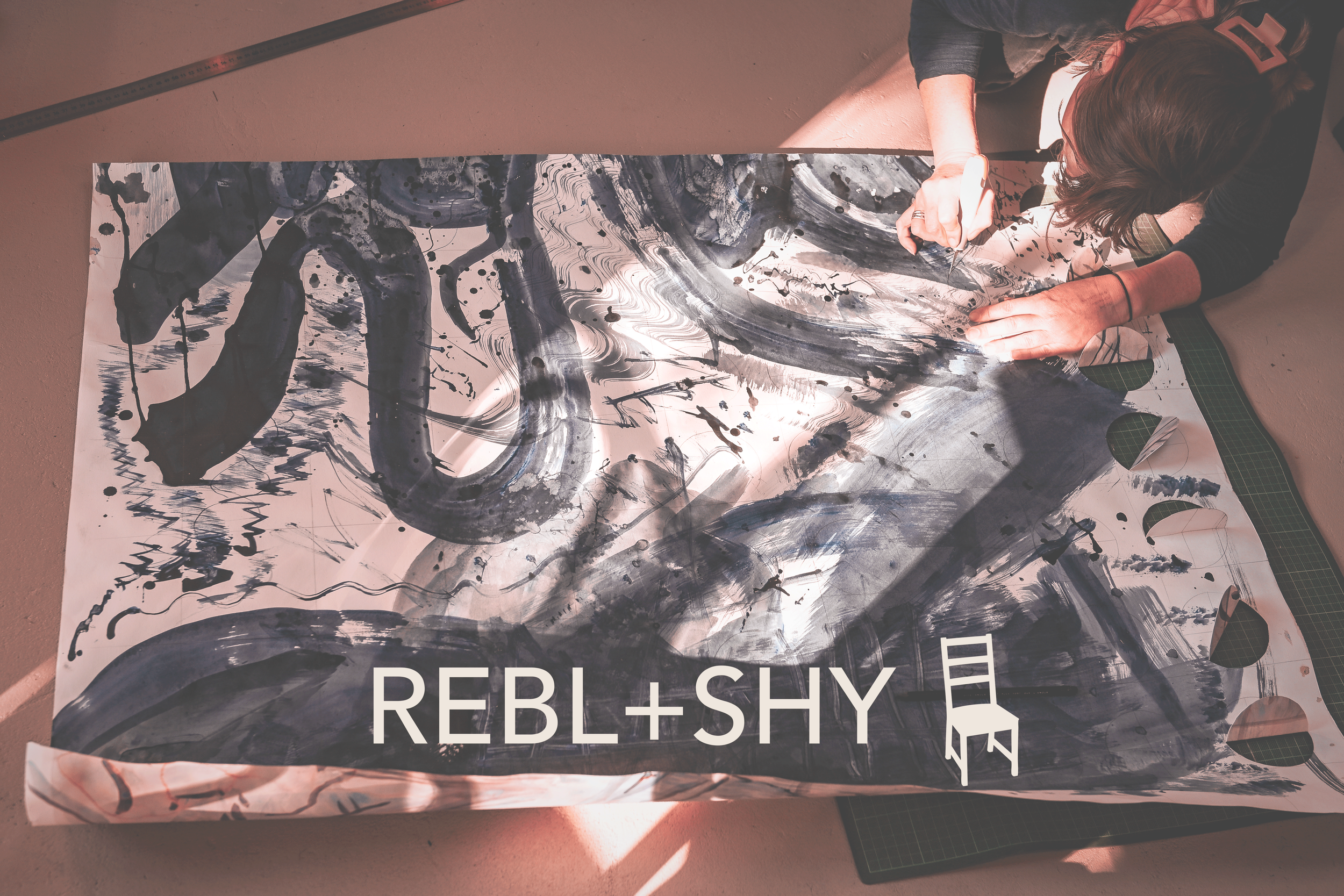 An artist works on a large piece of paper covered in various ink markings. The logo REBL+SHY is at the bottom of the image.