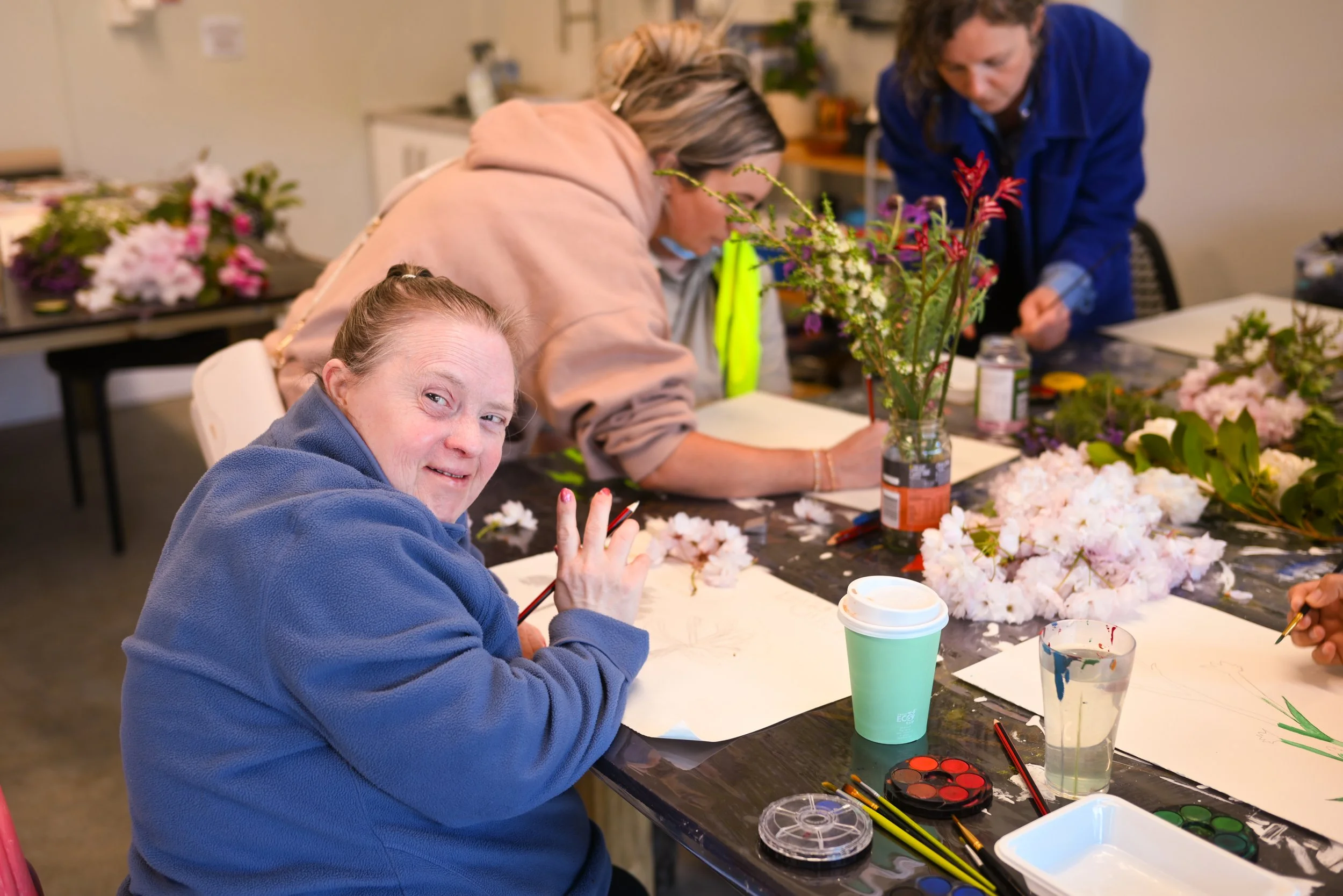 Artist Chrissie sits at a communal workspace and smiles and waves at the camera with a pencil in her hand. She has a piece of paper in front of her and the table has various spring blossoms on it. Two artists work in the background.