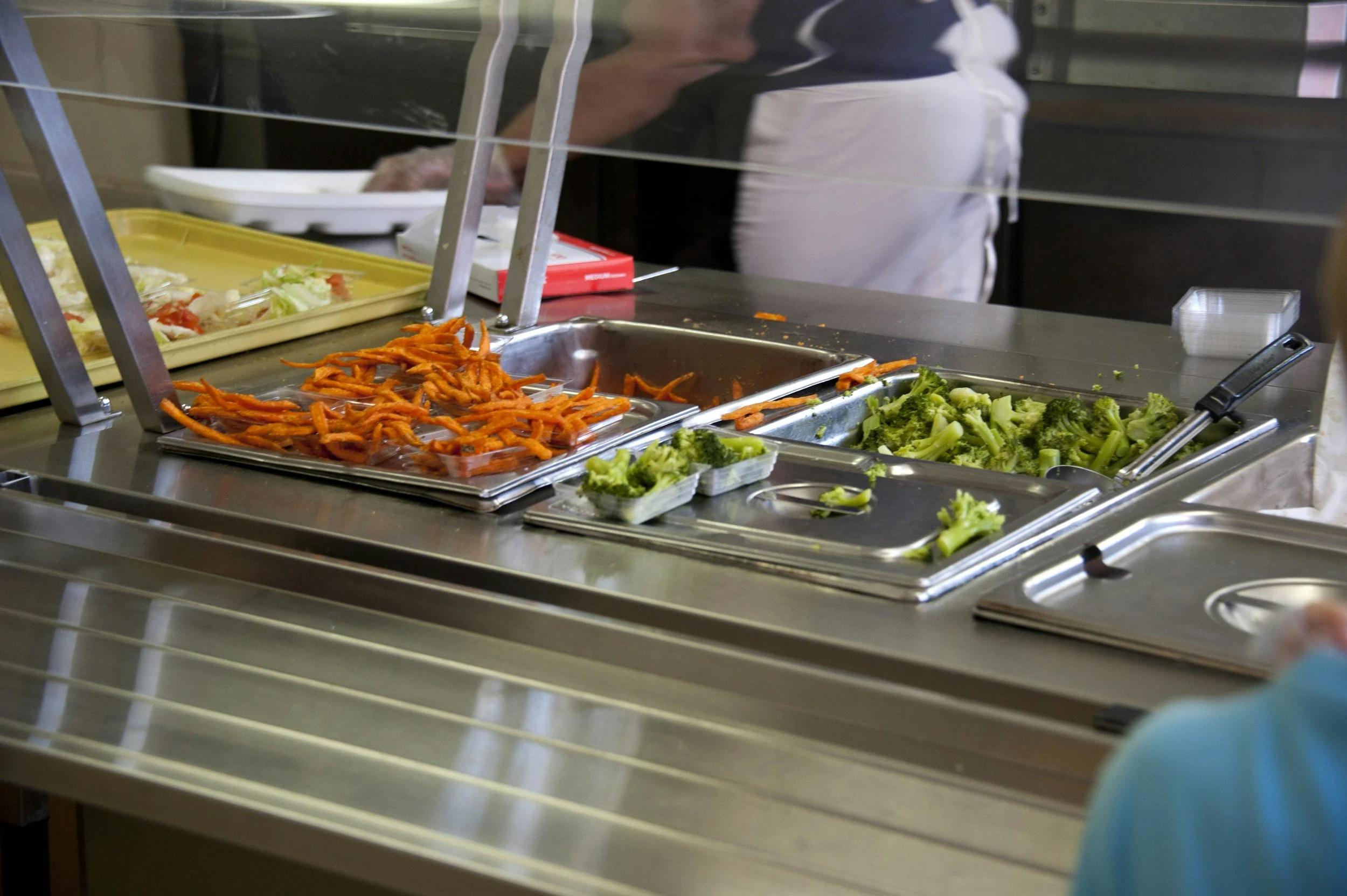 Salad bar with sliced carrots and broccoli on trays and trays of other vegetables behind a glass sneeze guard.