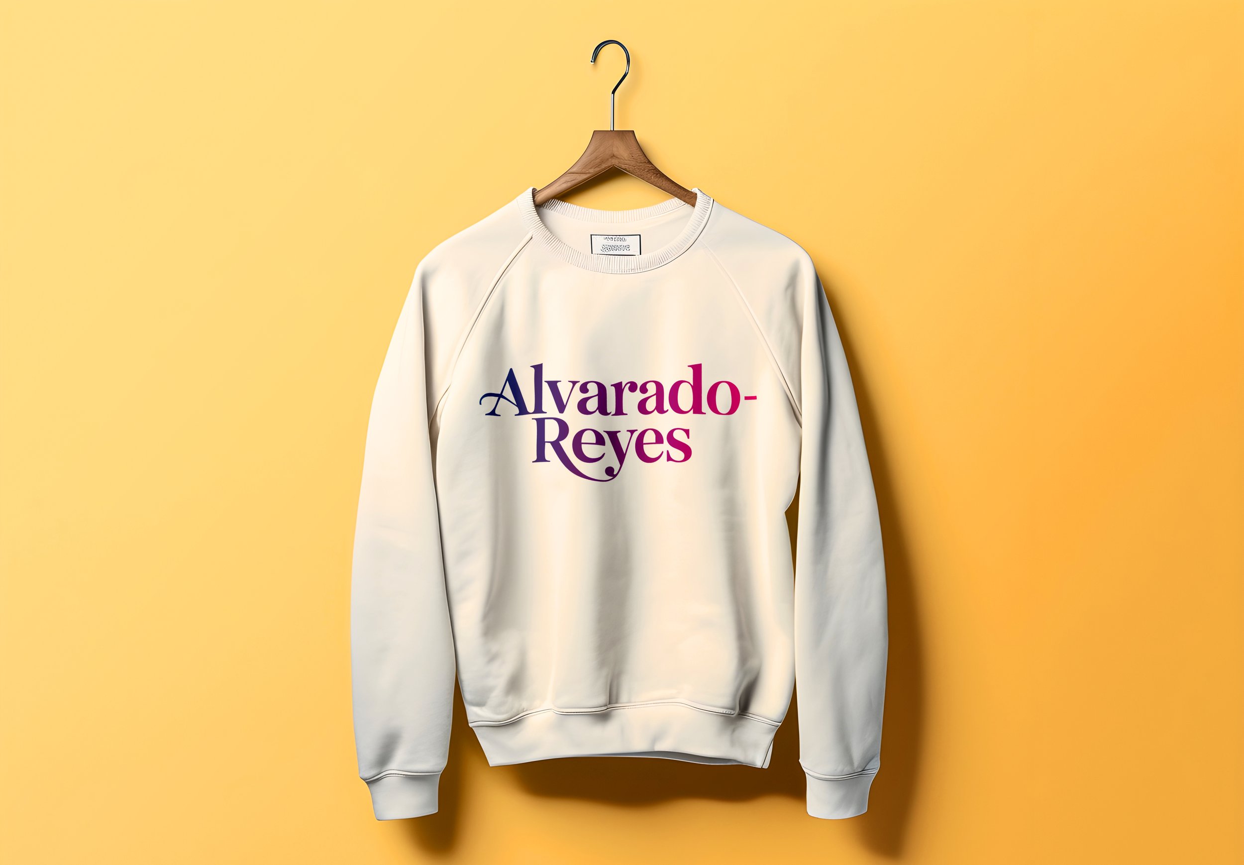 White sweatshirt with 'Alvarado-Reyes' printed in purple and pink letters, hanging on a wooden hanger against a yellow background.