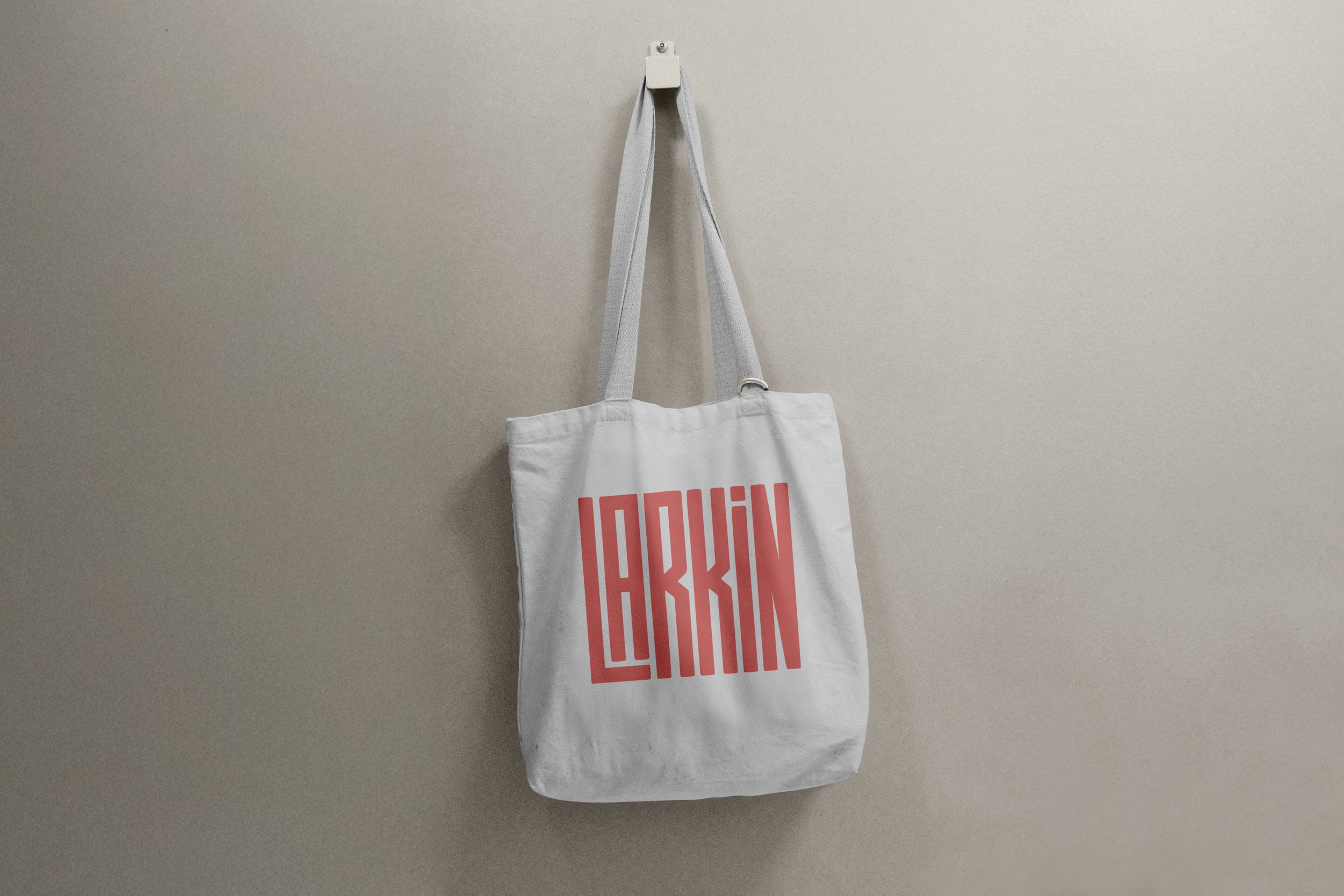 A white tote bag hanging on a wall hook against a plain, light-colored wall. The bag has the word 'LARKIN' printed in large, bold, pink letters.