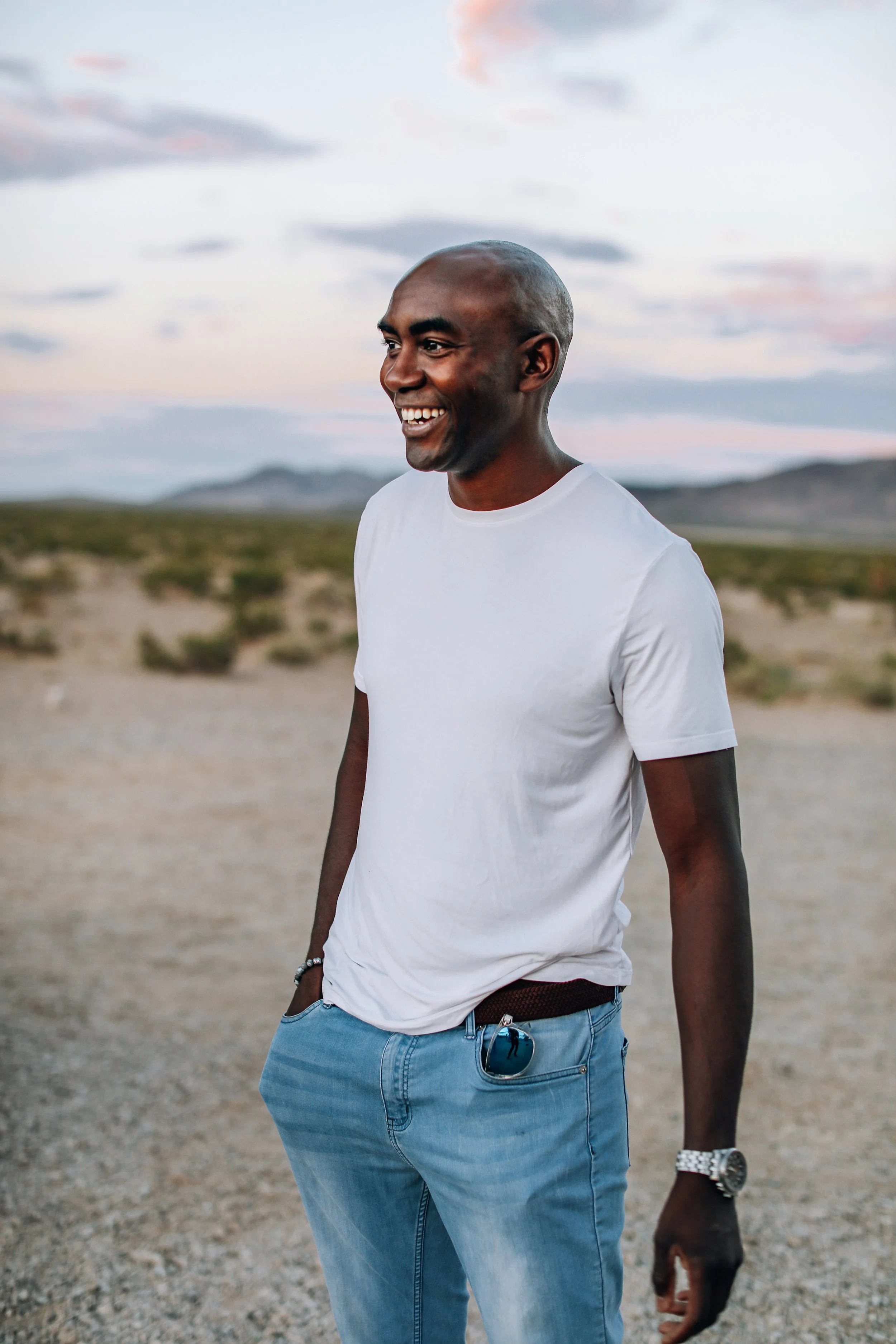 A smiling man wearing a white t-shirt, jeans, sunglasses in his pocket, and a watch, standing outdoors in a desert landscape during sunset.