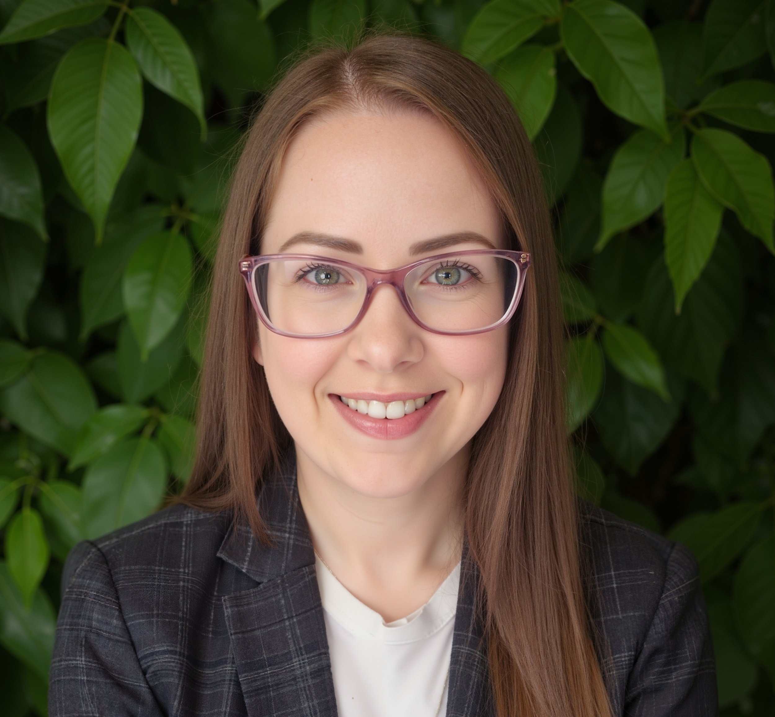 A woman with long brown hair, wearing light pink glasses, smiling, with green leafy plants in the background.