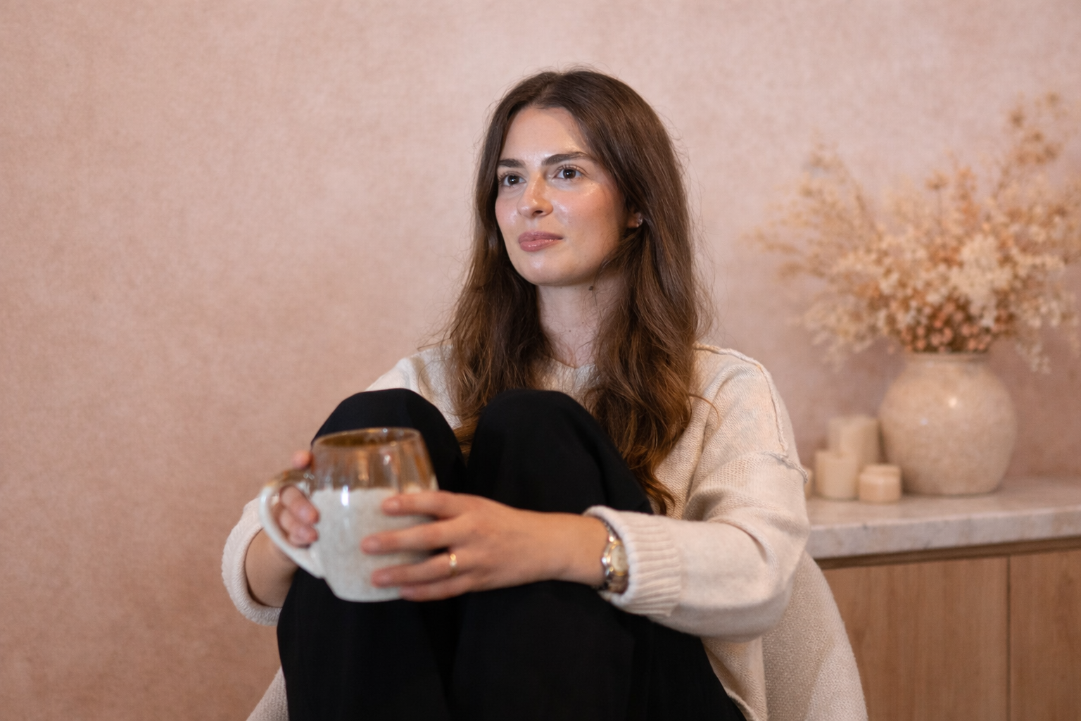 A woman with long brown hair sitting indoors, holding a ceramic mug, with a neutral expression. In the background, there is a vase with dried flowers and candles.