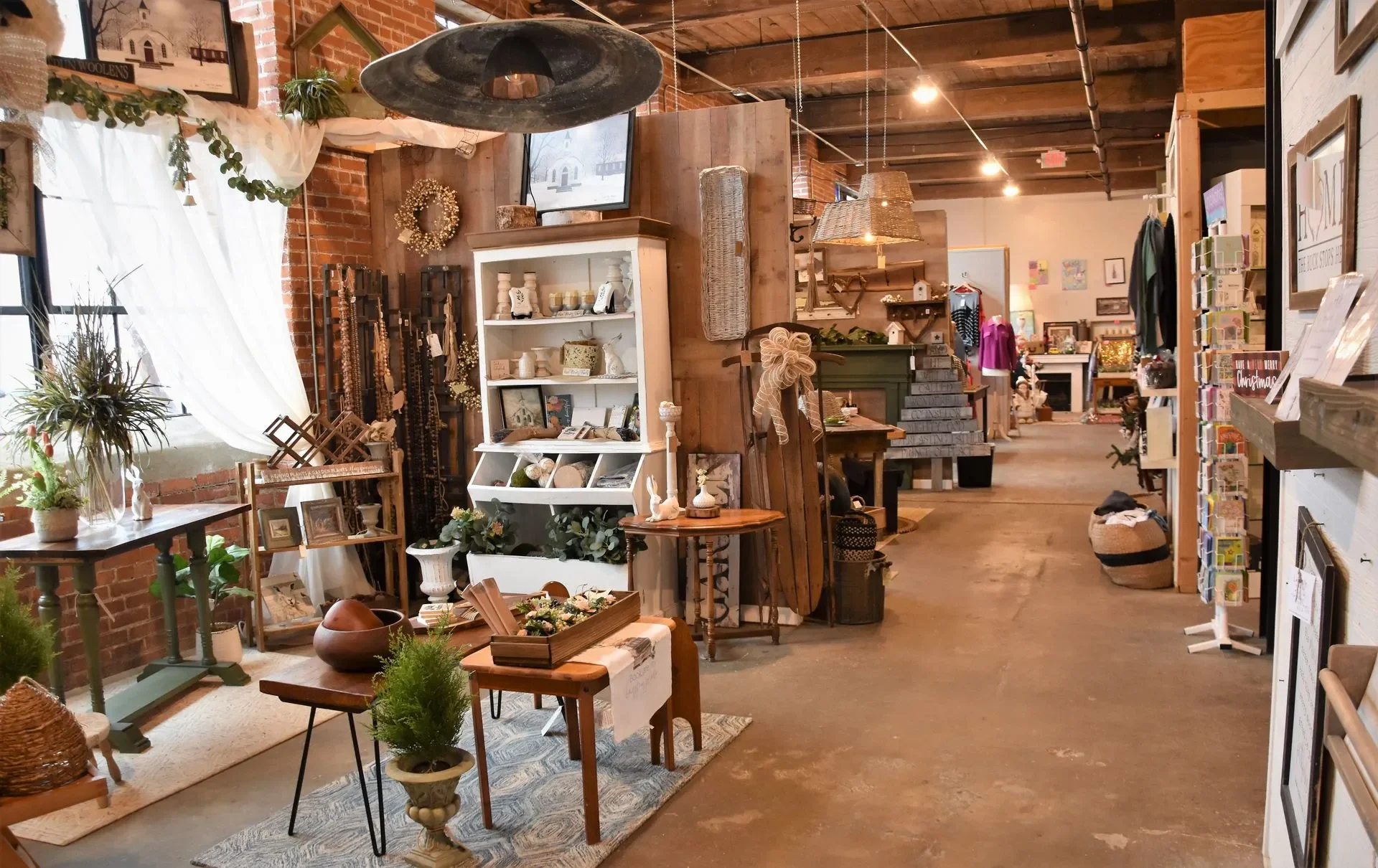 Interior of a boutique store featuring rustic decor, shelves with plants, jewelry, and home decor items, with a brick wall and curtains on the left, and various display racks and furniture throughout the space.