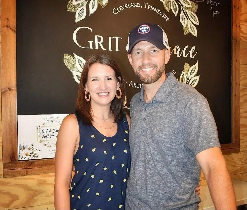 A man and woman smiling and posing together indoors, with a blackboard behind them that has white and gold lettering and decorative leaves, indicating Cleveland, Tennessee, with the words 'Grit and Grace' and 'Artisan' visible.