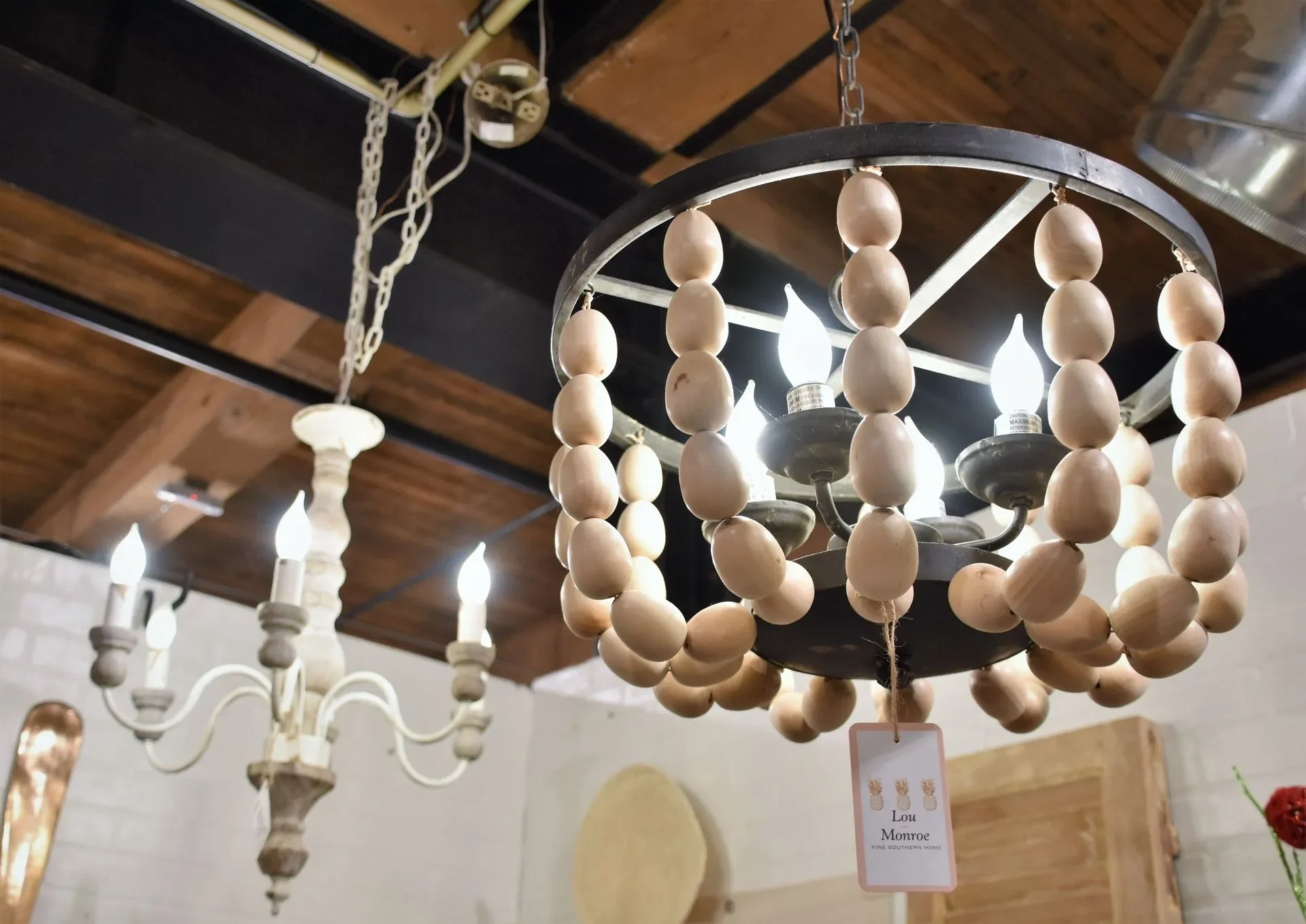 A chandelier with a metal frame and wooden beads surrounding the light bulbs, hanging from the ceiling with chains. In the background, there is another chandelier with candle-style lights.