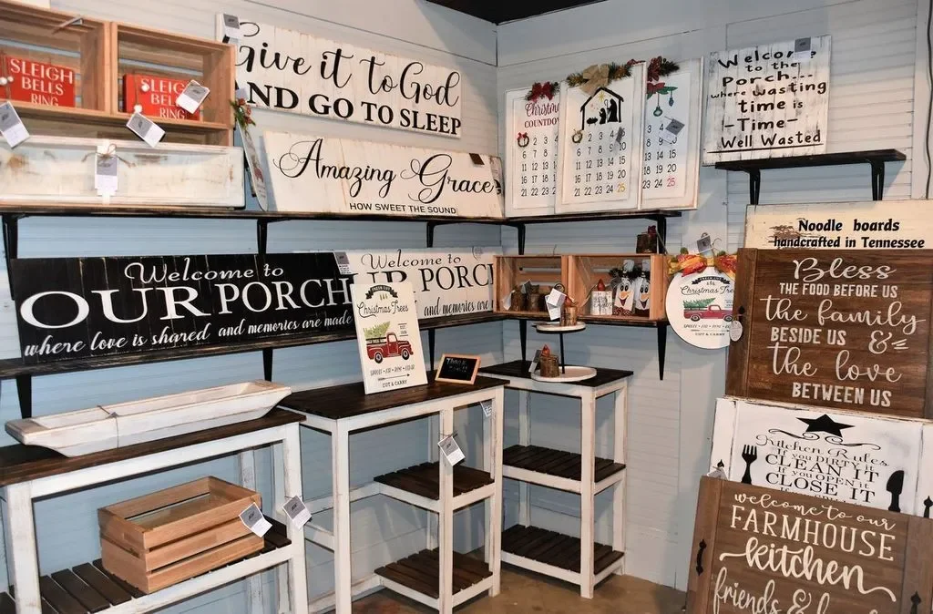 Shelf displays decorative wooden signs with inspirational quotes, Christmas themed items, and farmhouse style decor in a boutique store.