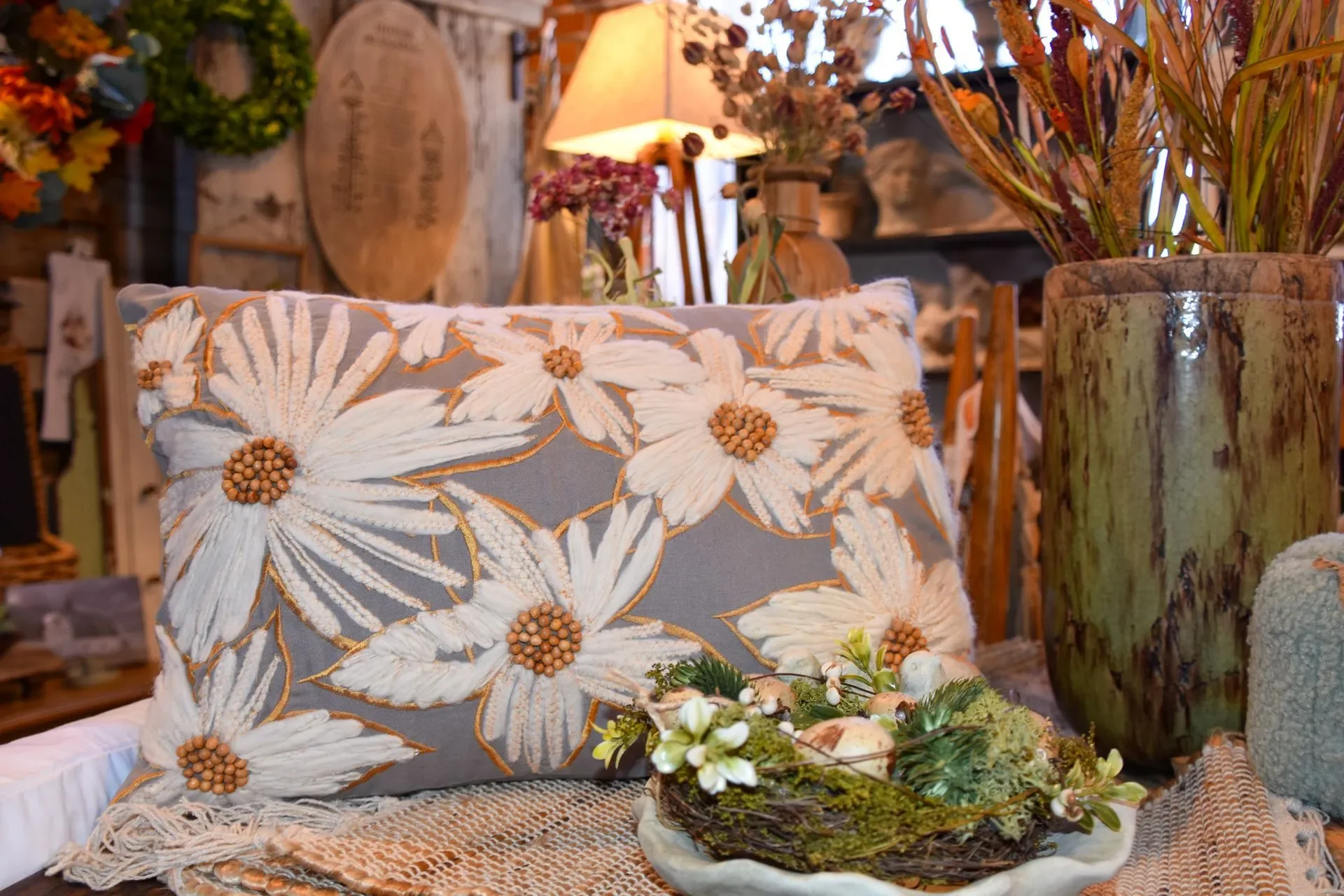Decorative beige pillow with embroidered white flowers and brown centers, surrounded by vases with dried flowers and a natural centerpiece, in a cozy interior setting.