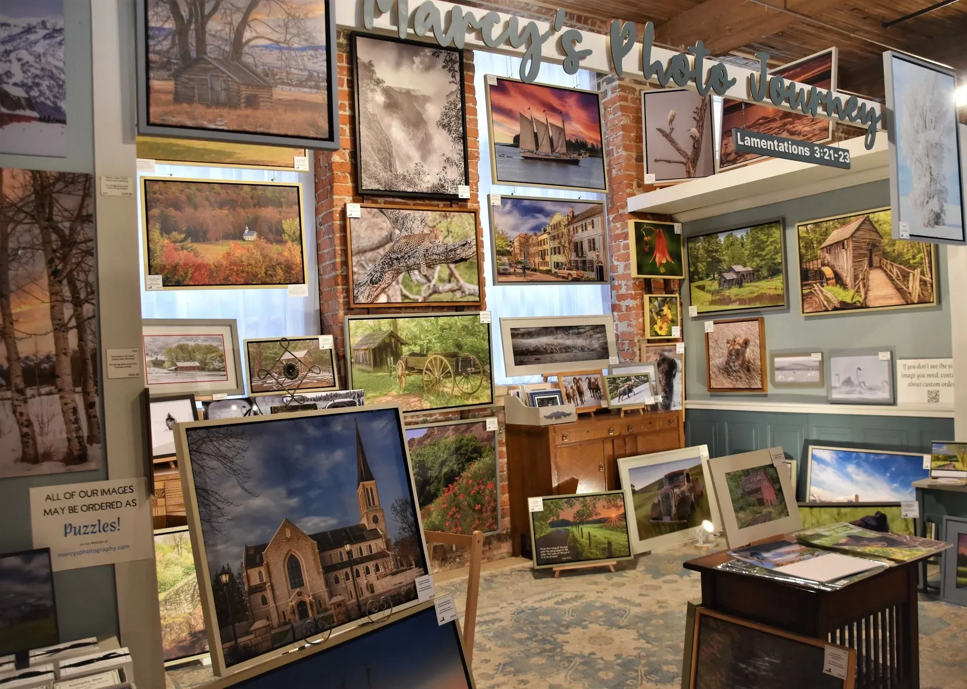 A gallery display with various landscape photographs, including mountains, forests, animals, and buildings, arranged on walls and easels in a cozy indoor setting.