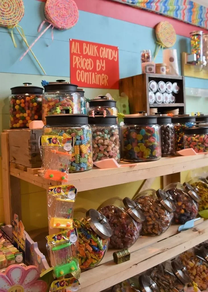 A candy store shelf with multiple large jars filled with colorful candies and chocolates. A red sign reads 'All bulk candy priced by container.' Pepperoni and other candies are visible in plastic containers on the lower shelf.
