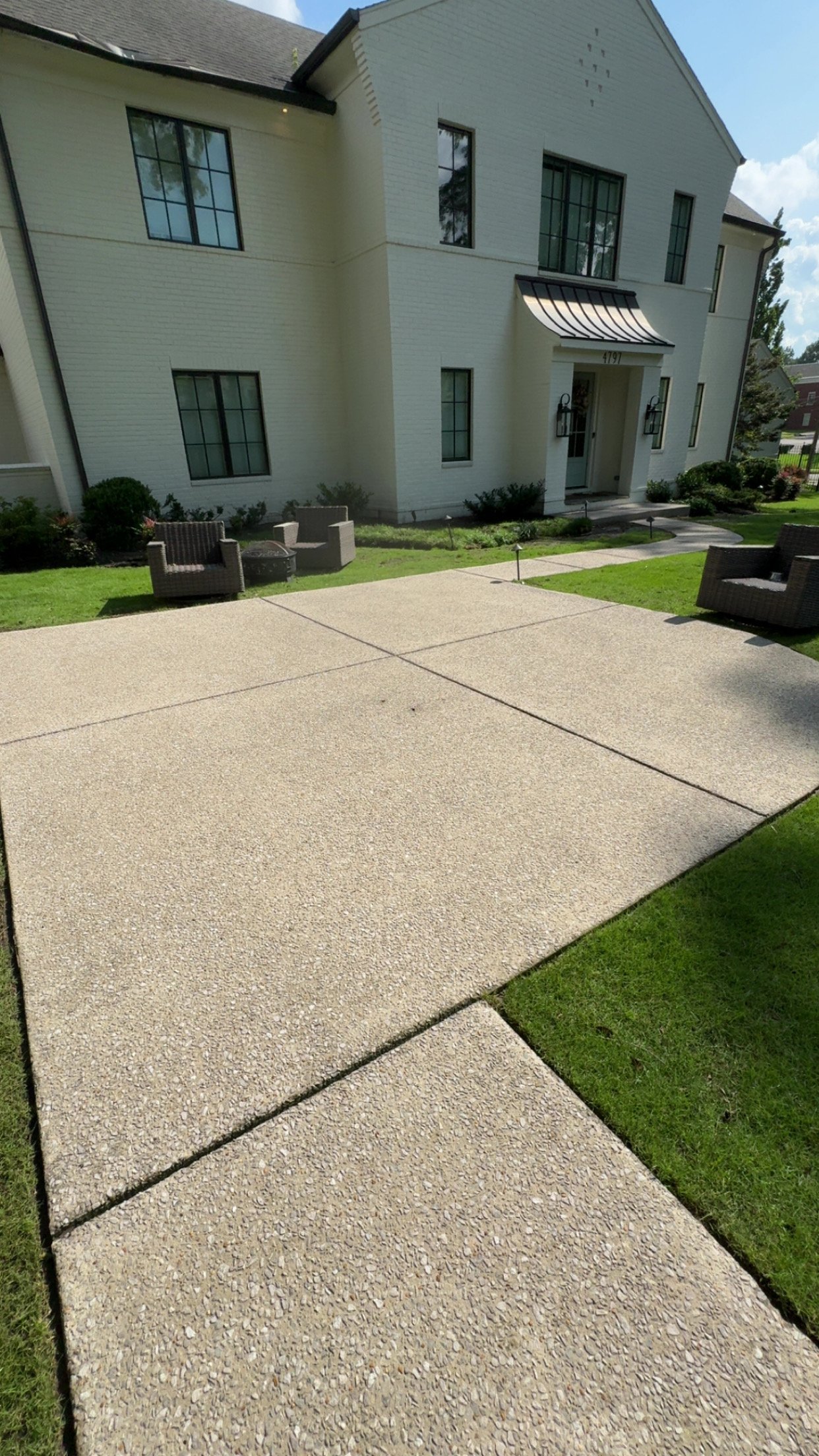 Concrete patio with grass edges in front of a white two-story house with multiple windows and an entry door with black lanterns.