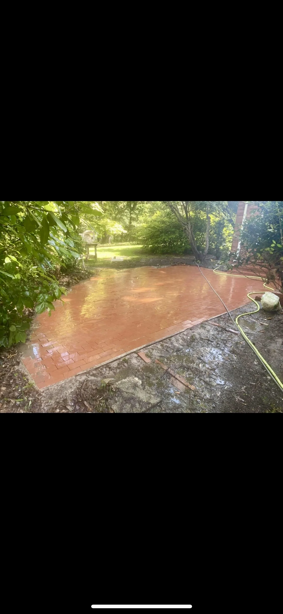 A brick pathway being washed with a hose, surrounded by greenery and trees.