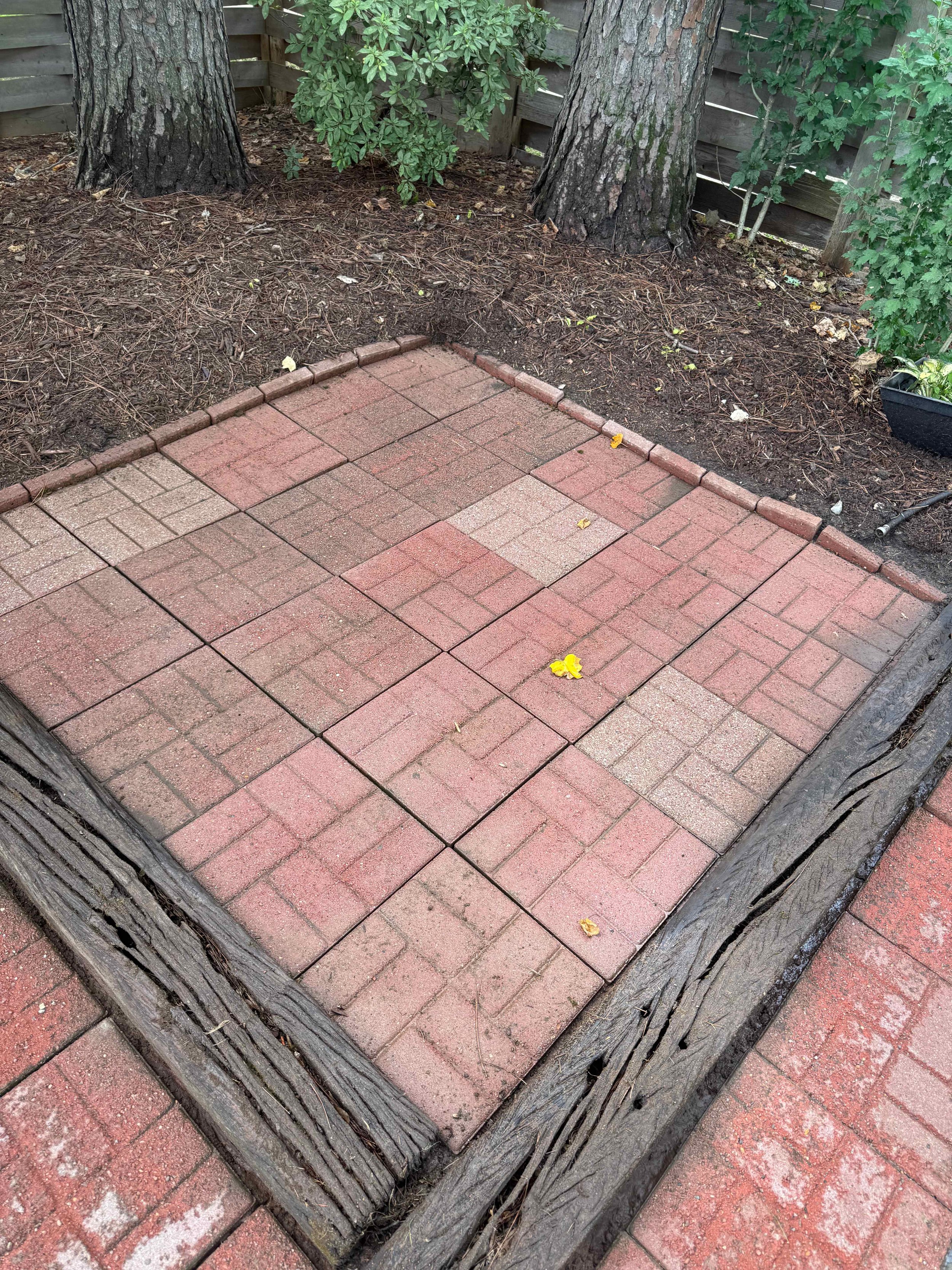 Newly installed brick patio with a border of old wooden railroad ties, surrounded by dirt, tree trunks, and garden plants.