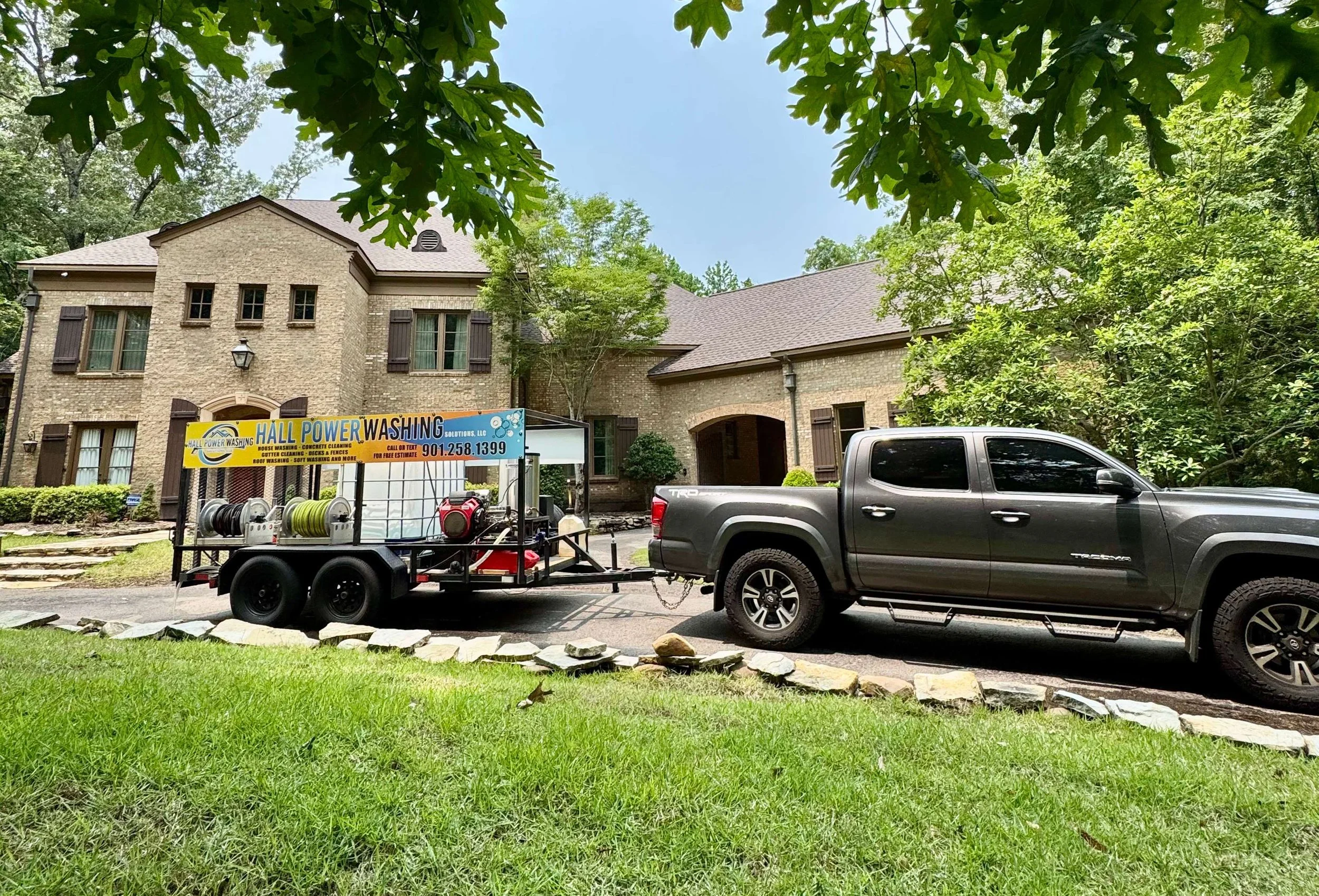 A black pickup truck with a trailer attached, carrying cleaning equipment and hoses, parked in front of a large brick house surrounded by trees and greenery.