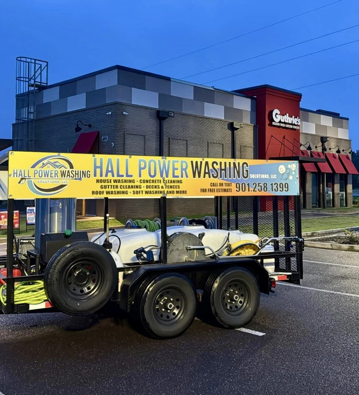 A power washing trailer parked in front of a building with Guthrie's chicken restaurant. The trailer has a bright yellow sign advertising various cleaning services and a phone number.