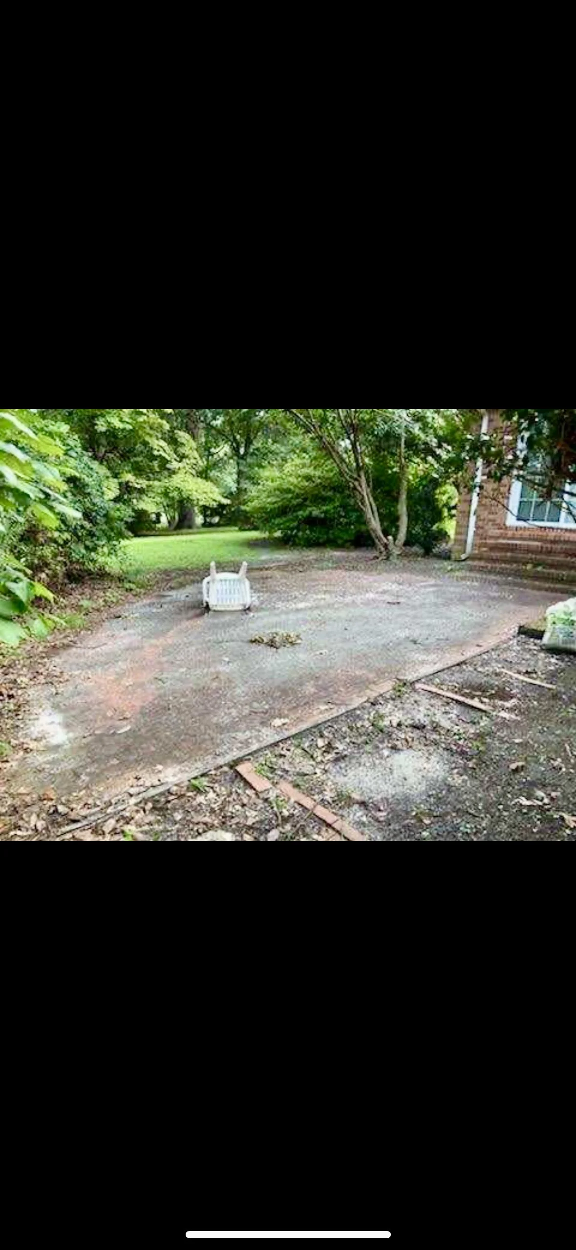 A backyard patio under renovation with bricks and dirt, a white plastic outdoor furniture piece on its side, surrounded by green trees and shrubbery, and part of a brick house visible on the right.