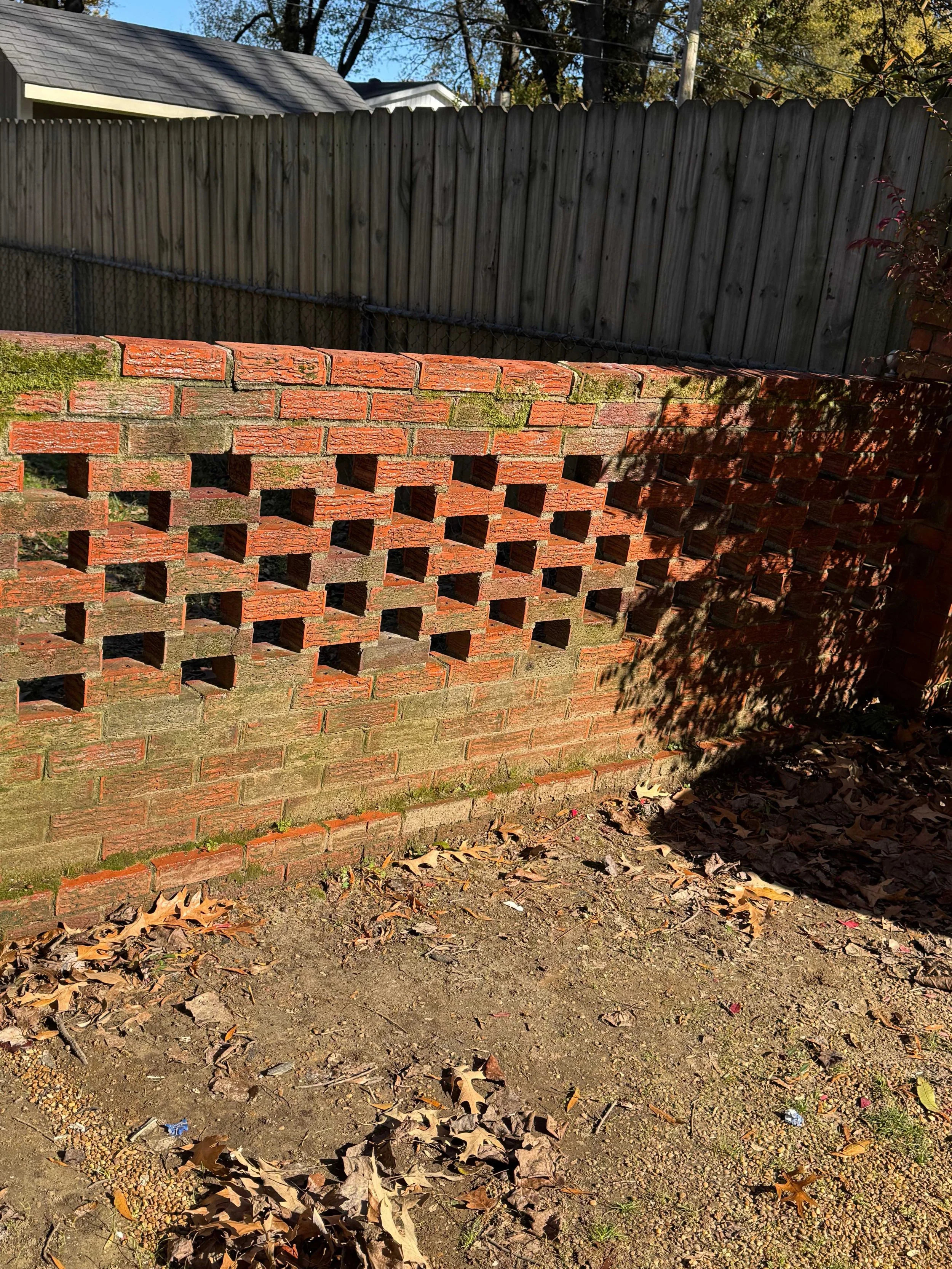 A red brick wall with square holes, partially covered with moss, with a wooden fence and trees in the background.