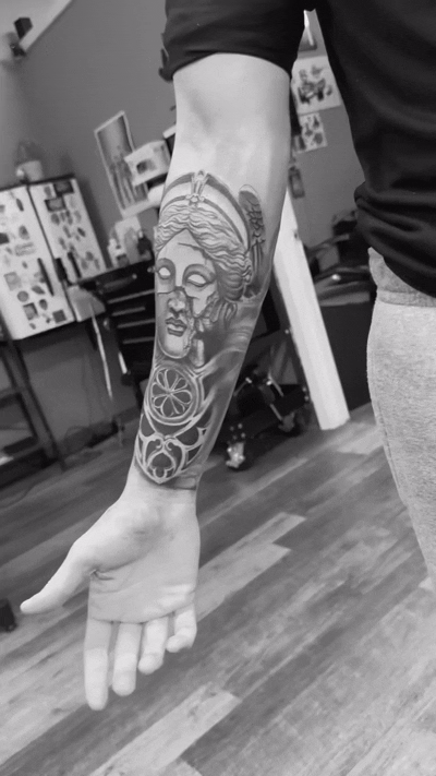 Black and white photo of a person's forearm with a detailed tattoo of a Greek goddess and geometric patterns.