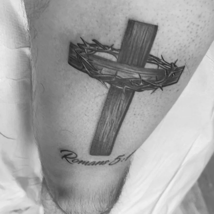 Tattoo of a wooden cross with a crown of thorns and Roman 5:8 written below.