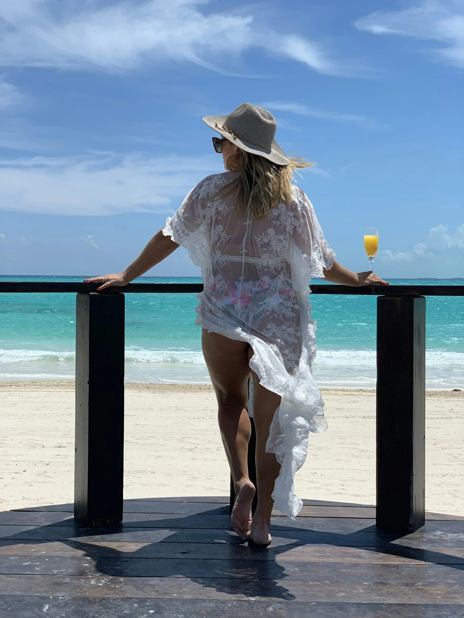A woman standing on a beach deck, wearing a sun hat, sunglasses, and a sheer white cover-up, looking out at the ocean while holding a glass of orange juice.