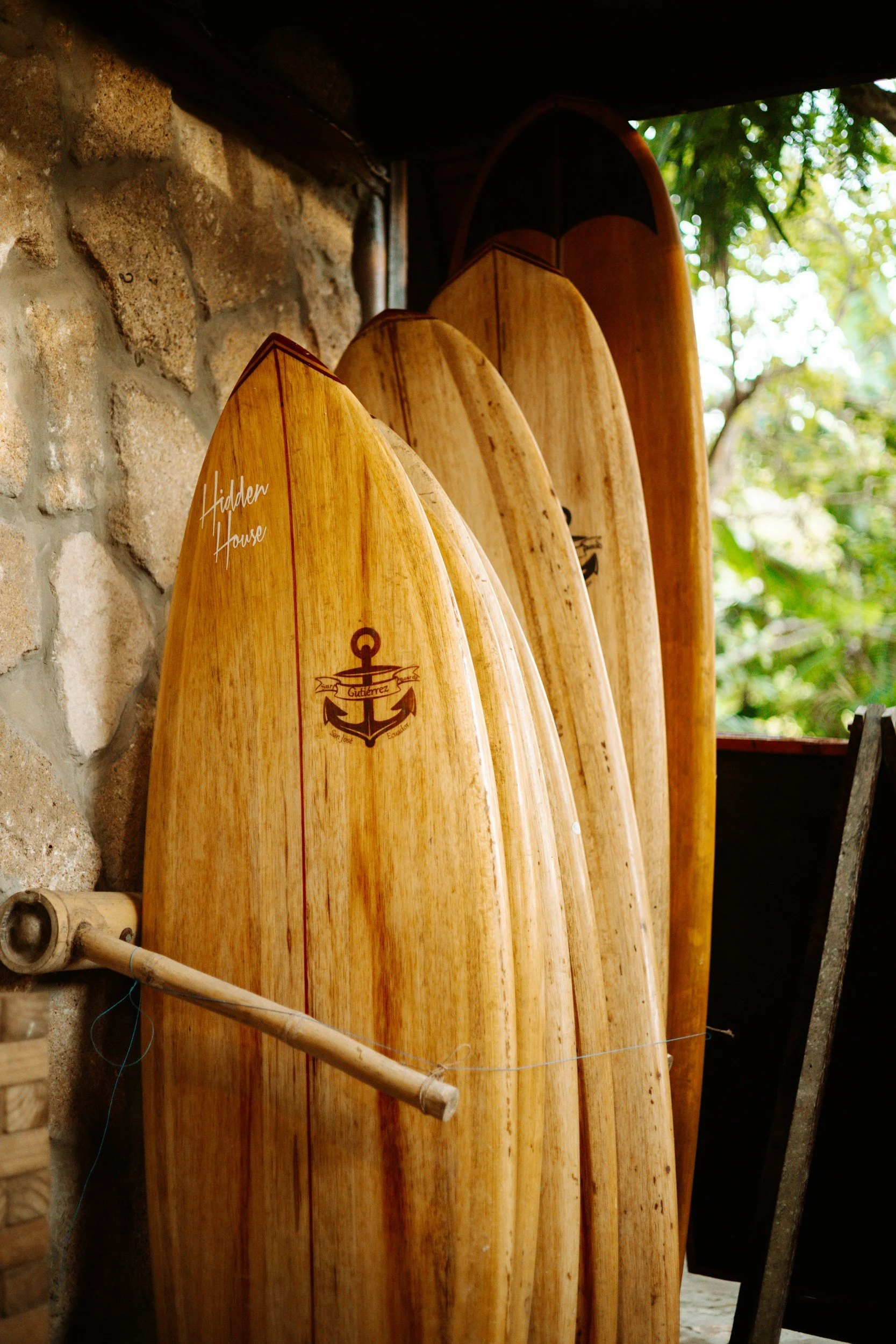 Multiple wooden surfboards leaning against a stone wall, with green foliage visible in the background.
