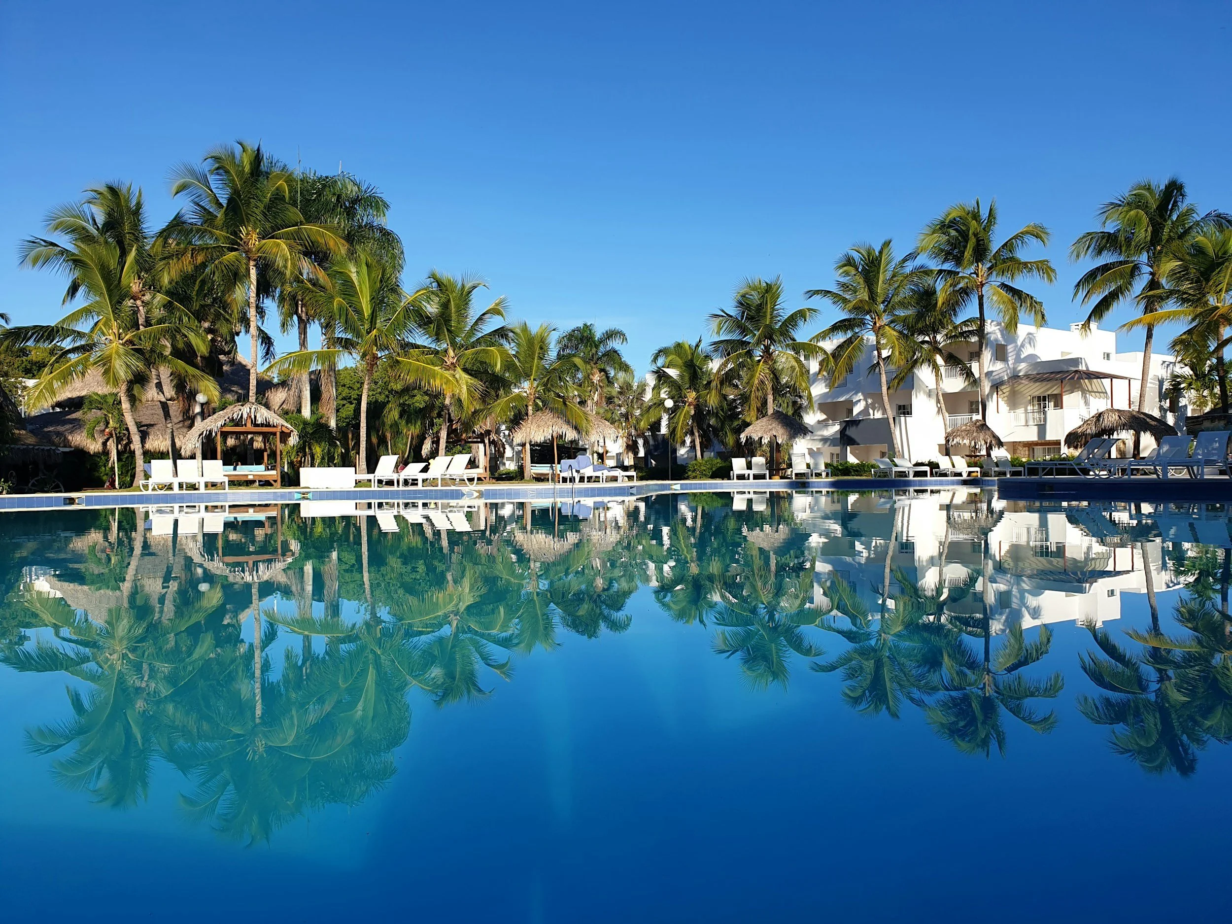 Resort pool area with clear water reflecting palm trees, white buildings, and thatched umbrellas under a blue sky.
