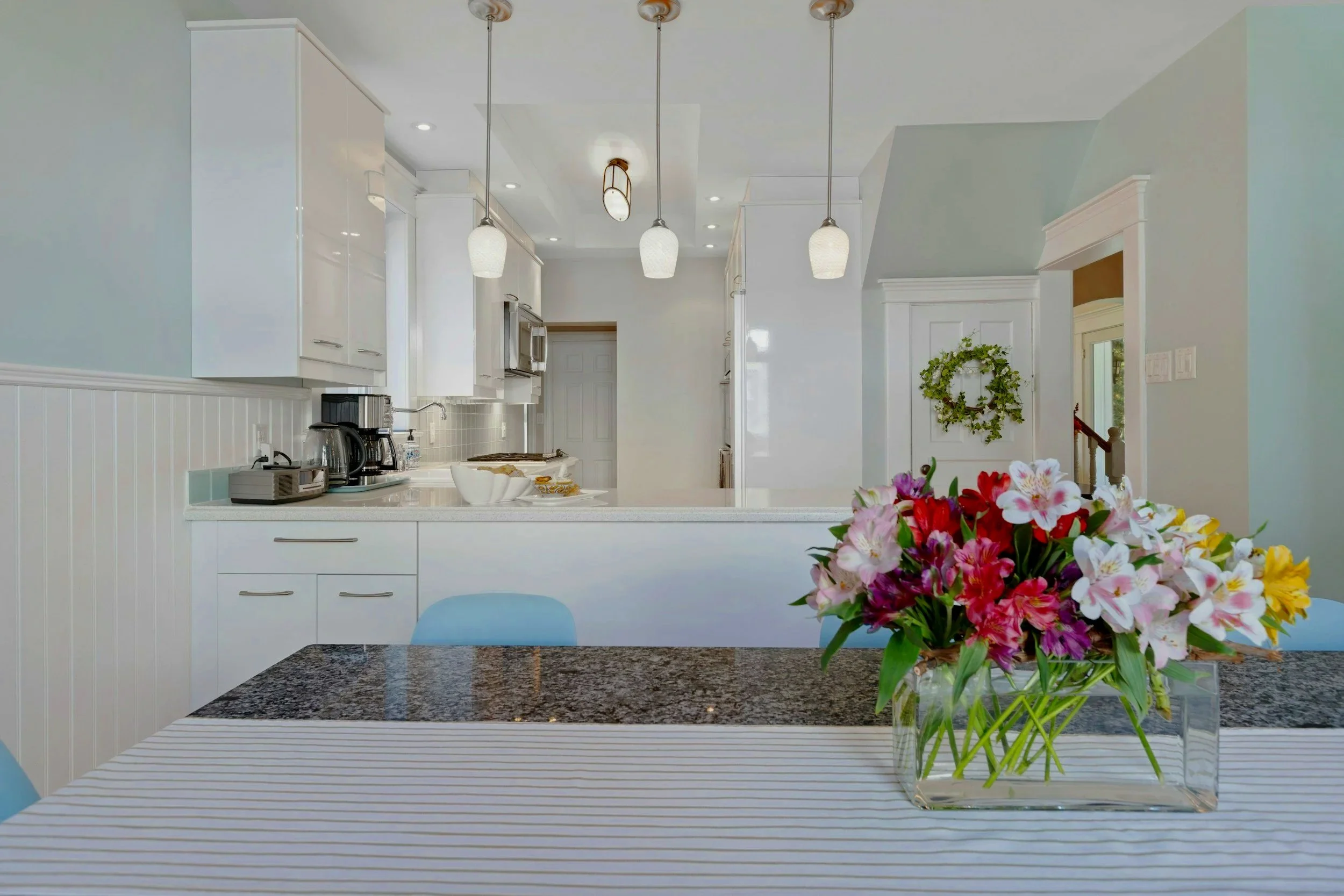 A dining table with a striped tablecloth and a glass vase of pink, white, purple, and yellow flowers. In the background, a modern white kitchen with pendant lights, white cabinets, and some appliances.