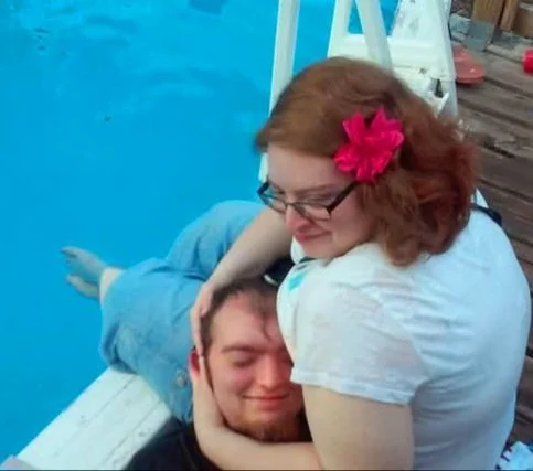 A woman with red hair and glasses holding and embracing a smiling young man with closed eyes.