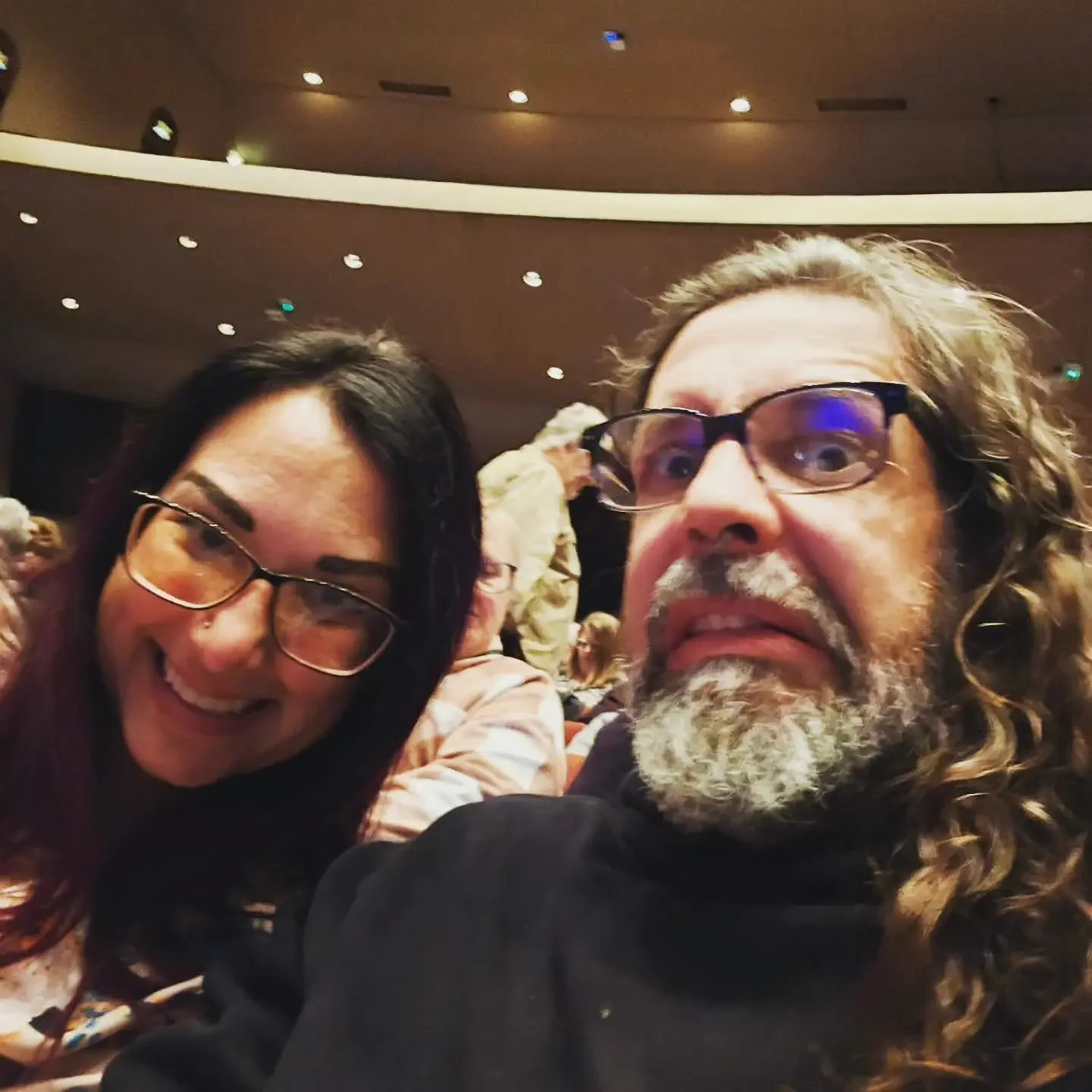 Close-up selfie of a woman with long dark hair, glasses, and a big smile, and a man with curly long hair, a beard, glasses, and a surprised expression, at an indoor event with a crowd in the background.