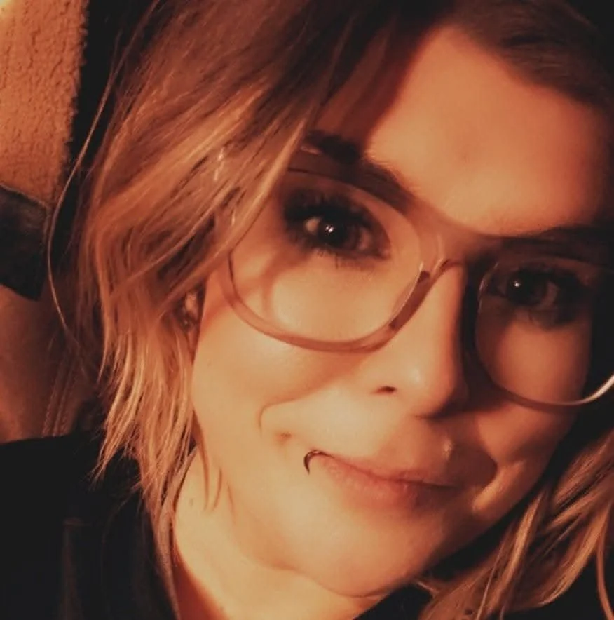 Close-up of a young woman with light brown hair, wearing glasses, smiling with a nose piercing, in a cozy indoor setting.