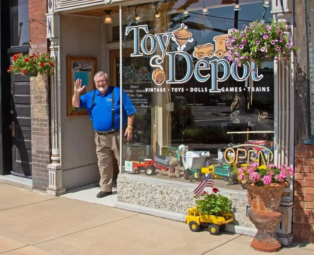 A man standing outside a toy store named Toy Depot, waving at the camera. The store's window displays toy trucks and other toys, with a sign indicating it is open. There are flower pots with pink and purple flowers on either side of the window and sidewalk, and a small yellow toy truck with a potted plant and American flag in front. The store sells vintage toys, dolls, games, and trains.