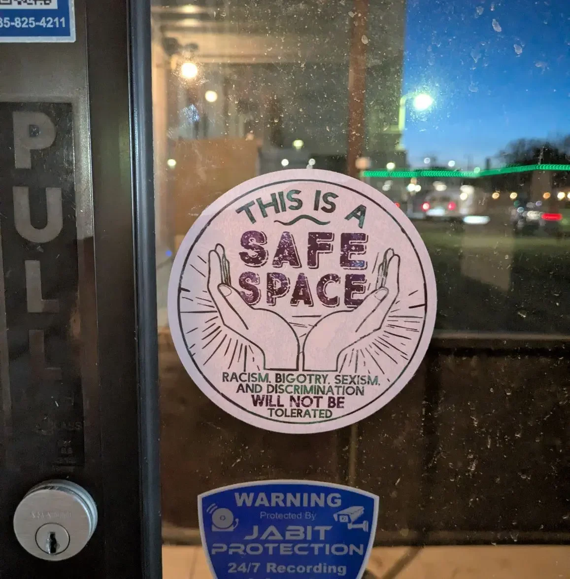A sticker on a door with the message: "This is a safe space. Racism, bigotry, sexism, and discrimination will not be tolerated," featuring two hands forming a protective gesture around the words. There is also a blue security sticker with the text: "Warning protected by JABIT PROTECTION 24/7 recording."