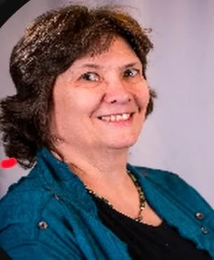 A woman with short brown hair smiling, wearing a teal jacket and a black top, with a beaded necklace.