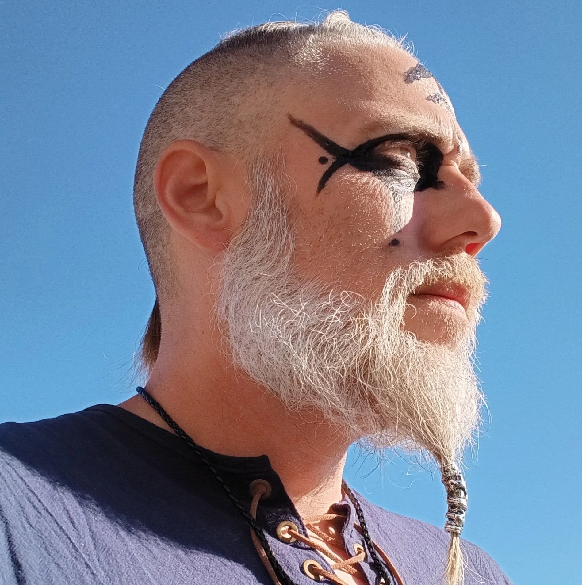 Close-up side view of a person's face with a styled beard and mustache, wearing face paint resembling a skull with black details around the eye and cheekbone, set against a clear blue sky.
