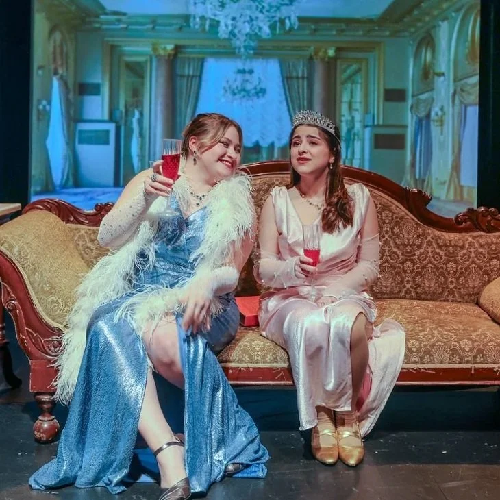 Two women sitting on an ornate couch, wearing elegant dresses and tiaras, holding glasses of red drink, in an opulent room with chandeliers and draped curtains.