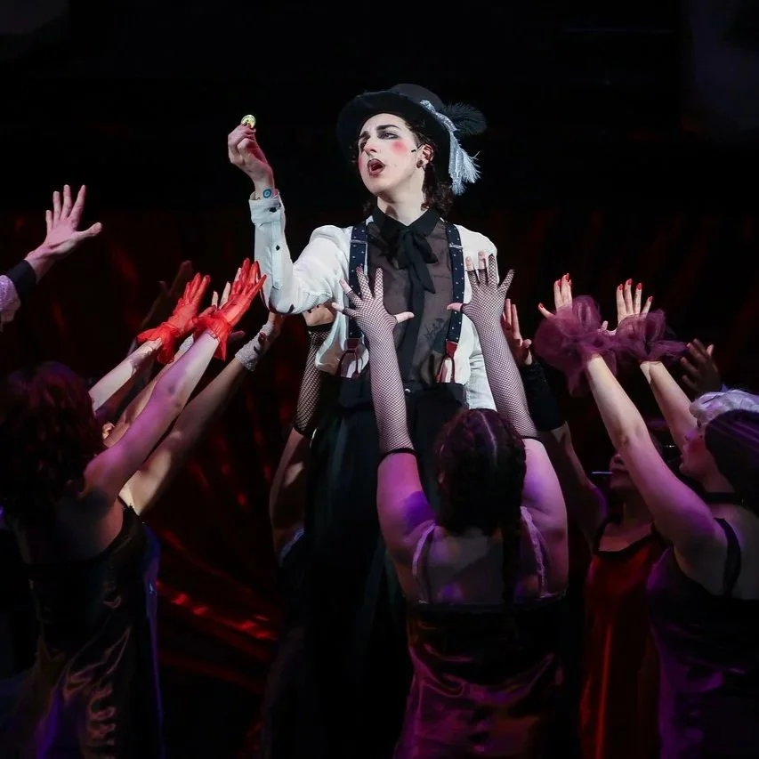 A woman dressed in vintage style with a black hat and white blouse stands on a stage, surrounded by performers reaching up towards her with hands wearing black fishnet gloves. The scene appears to be a theatrical performance.