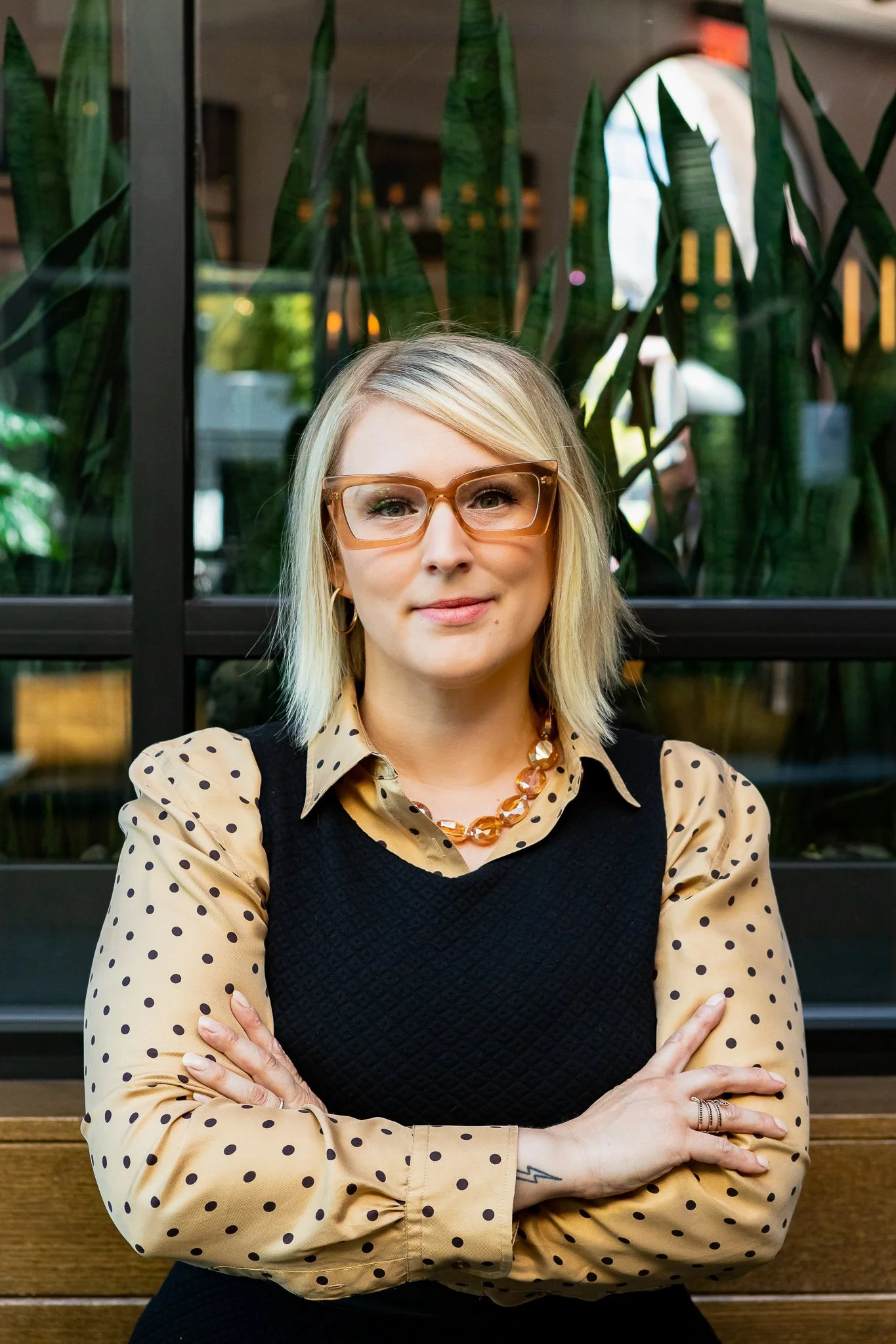 a blonde girl with unique glasses stands confidently with her arms crossed