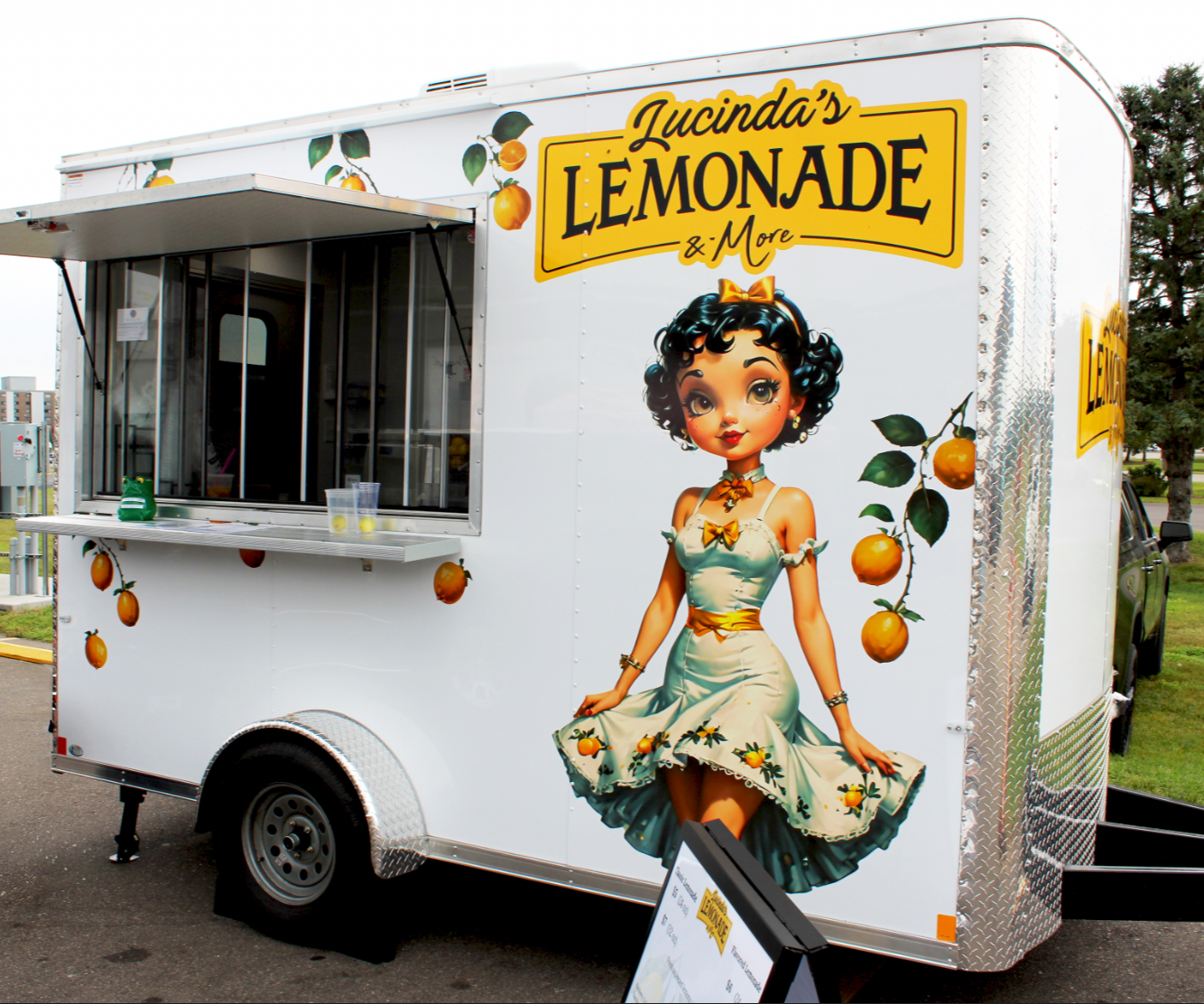 A white lemonade stand trailer featuring colorful graphics of a vintage-style cartoon girl with black, curly hair and a yellow bow, surrounded by lemons and lemon branches, with a sign that reads 'Lucinda's Lemonade & More'.