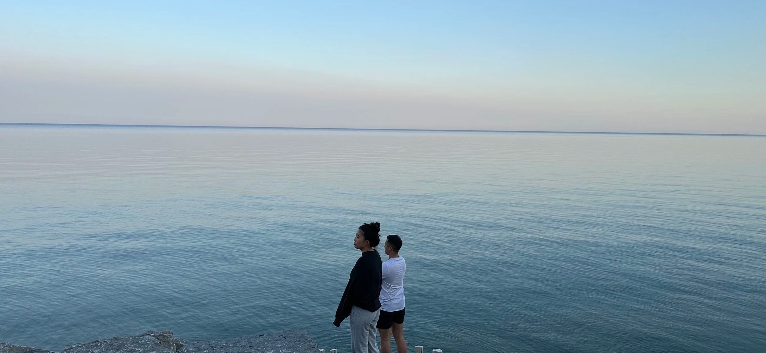 Two people standing together at the water's edge at sunset, looking out over calm water